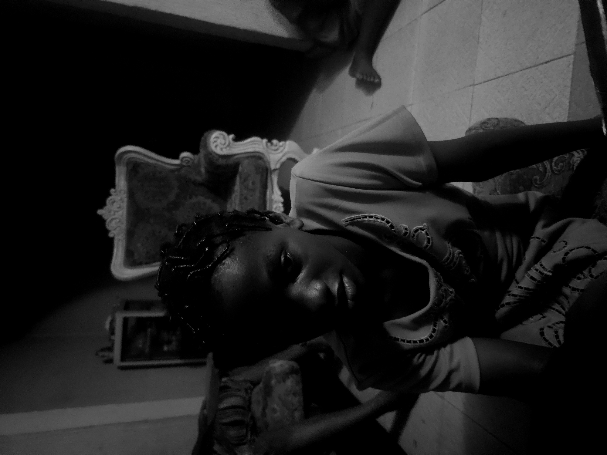 This black and white, vertically oriented photograph depicts a dimly lit indoor scene. The central subject is a person, likely a woman or young girl, whose face is partially illuminated by a light source, highlighting one eye, parts of her nose, and lips. Her hair appears to be styled in dark braids or twists. She is wearing a light-colored top with noticeable embroidery or patterns on the collar and sleeve. Her left arm is visible, resting in the foreground.
The setting is an interior space, likely a home. To the left and behind the person, an ornate piece of furniture, possibly a chair or sofa, with carved elements and patterned upholstery, can be seen. Further back on the left wall, a framed picture or mirror is visible. The floor is tiled. In the upper right quadrant of the image, a pair of bare feet is visible against the tiled surface, suggesting another person or the subject's own feet resting on an elevated surface.
The image captures a still moment; the person appears to be looking towards the viewer, possibly posing. The low-light conditions create strong contrasts and deep shadows, indicating it could be evening, nighttime, or a very dimly lit room. The person's expression is somewhat obscured but suggests a calm or contemplative mood. No visible text is present.