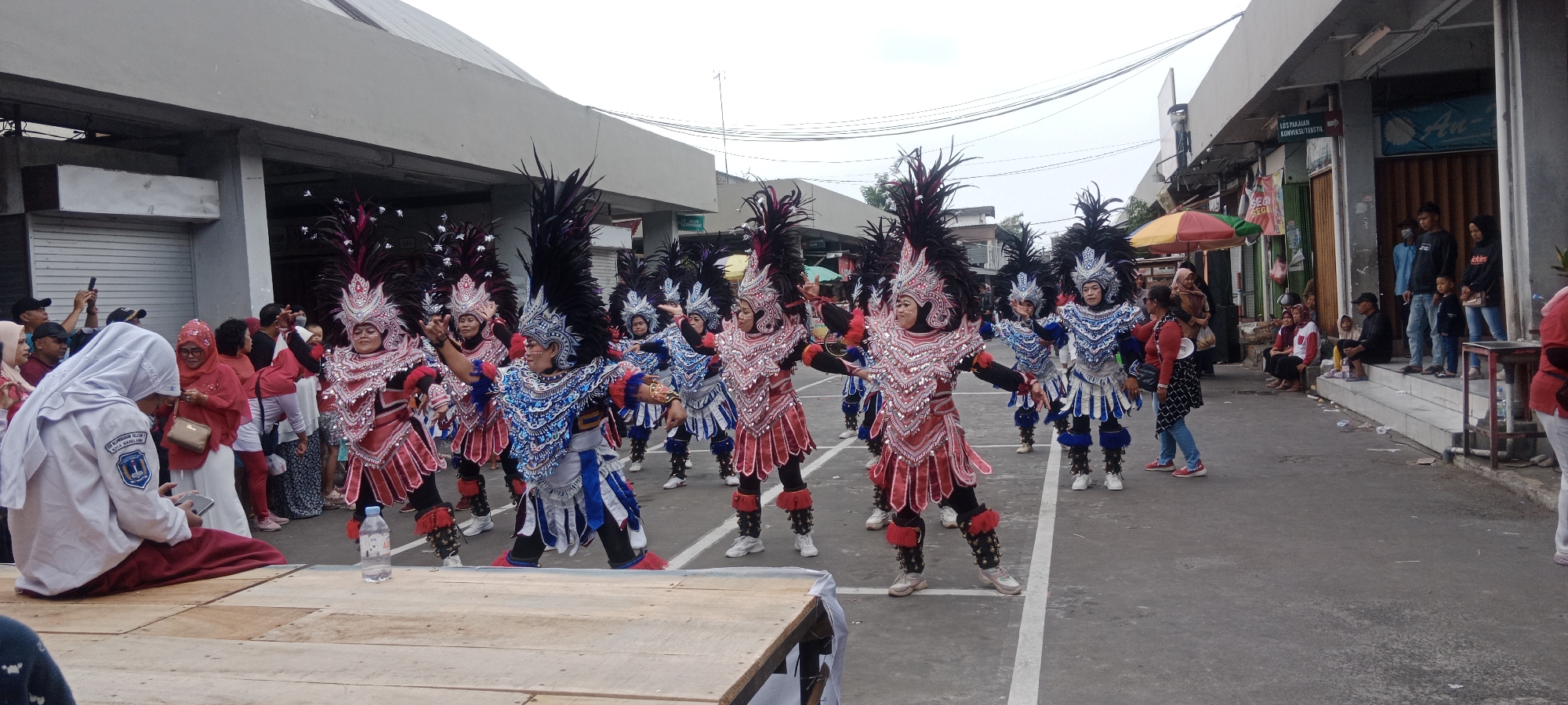 In the city of Magelang, Indonesia, a group of performers dressed in ornate, brightly colored costumes with large headdresses of black and red feathers are participating in a parade or cultural event. The costumes feature intricate detailing, including reflective elements and layered fabric, with some performers wearing red and silver ensembles and others in blue and silver. They are dancing or posing in the street, with several people in the foreground and background observing the spectacle.
On the left side of the image, a person in a white hijab and uniform is seated on a wooden platform, looking at a smartphone. Beside them, other spectators, mostly women in hijabs and traditional clothing, are watching the performers. A bottle of water is placed on the wooden platform.
The background shows a street lined with buildings that appear to be shops or market stalls, with awnings and signage visible. Some of these buildings are closed with metal shutters. Several people are seen standing or sitting outside these establishments, observing the parade. The sky is overcast, suggesting it might be a cloudy day. The scene captures a moment of cultural celebration or public performance within an urban marketplace setting in Magelang.