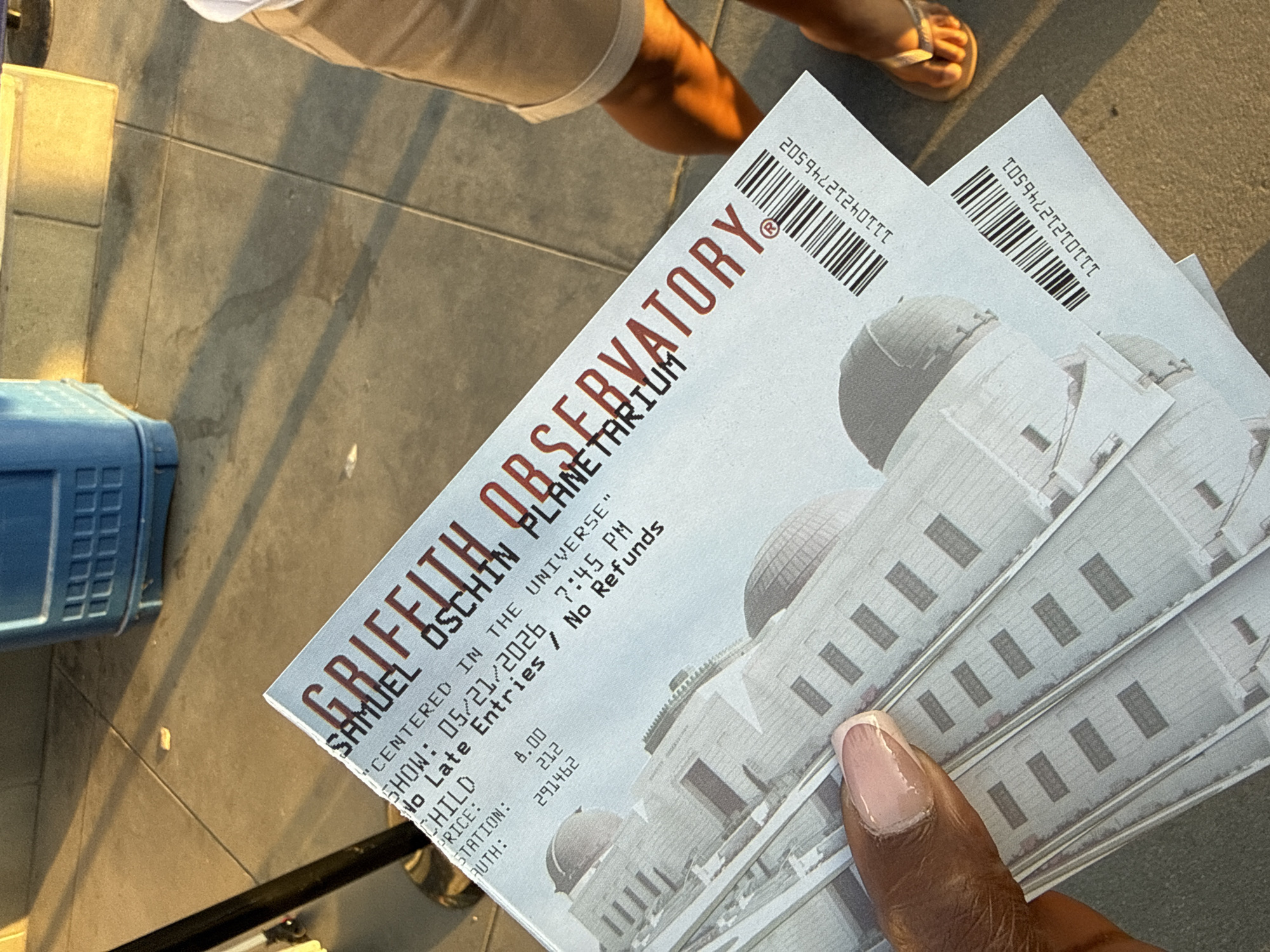 An eye-level outdoor shot shows a hand holding several tickets to the Griffith Observatory’s Samuel Oschin Planetarium in Los Angeles. The tickets feature a picture of the white art deco observatory. Text on the ticket includes show date (05/21/2026), time (7:45 PM), and the price for a child’s ticket ($8.00). Above the tickets is a partial view of two light-skinned people wearing sandals. A blue object is located to the left of the tickets. The background consists of pavement.