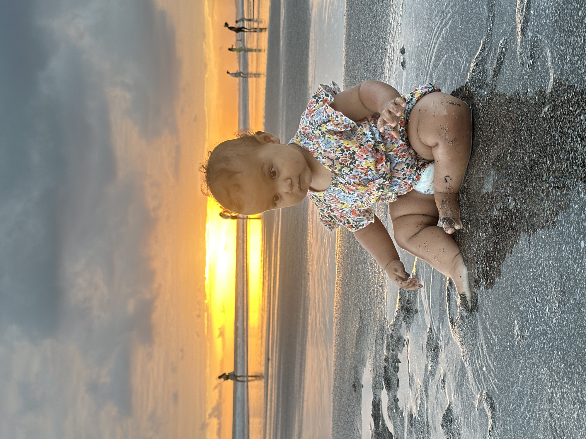 A young child is sitting on a sandy beach in North Kuta, Indonesia, during sunset. The child is wearing a floral-print onesie and has sand on their legs and feet. In the background, the sun is setting over the ocean, casting an orange and yellow glow on the water and sky. Several other people are visible in the distance, walking along the shoreline. The tide is coming in, with shallow water covering parts of the sand around the child.