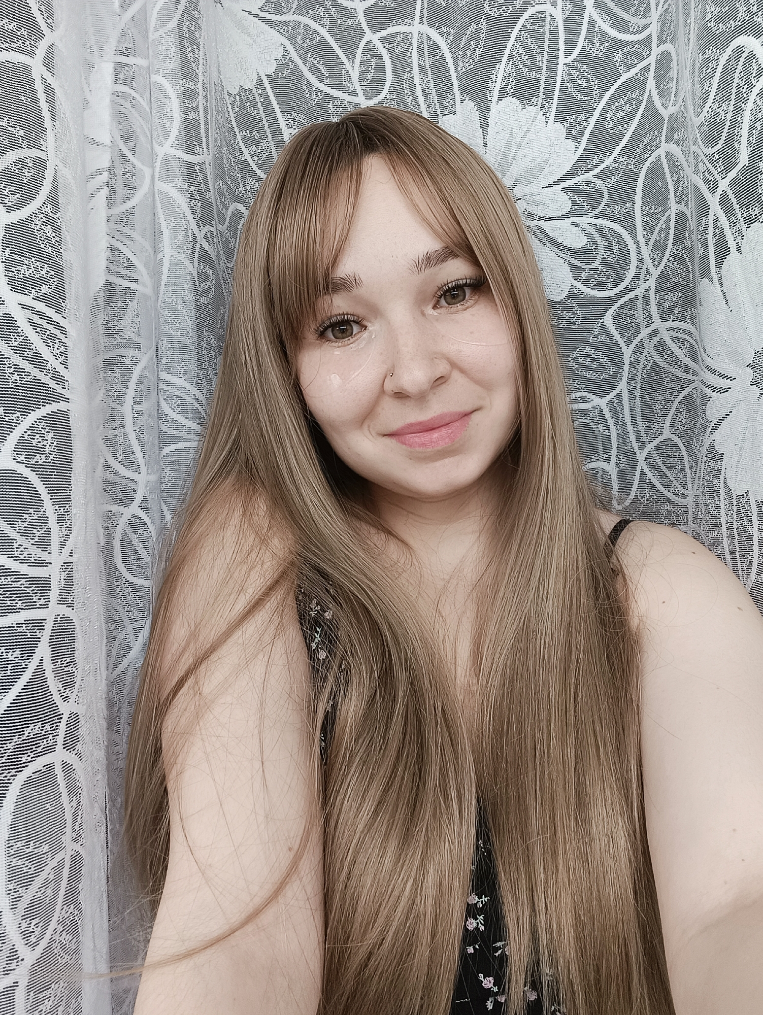 A headshot-style image displays a single young adult female looking directly at the camera. She possesses a light complexion, long, straight, light brown hair with bangs, and light brown eyes. A small silver ring is visible in her left nostril. Two translucent, possibly hydrogel, patches are applied to the skin directly beneath each eye. Her lips are slightly parted in a smile, exhibiting a light pink tint. She is dressed in a dark-colored garment featuring a small, light floral pattern of white and pink flowers, with a black spaghetti strap visible on her right shoulder. The background consists of white lace curtains, characterized by a repeating pattern of stylized floral and leaf motifs, against a dark, featureless backdrop. The specific location within Izhevsk, Russia, is not ascertainable from the visual information presented.