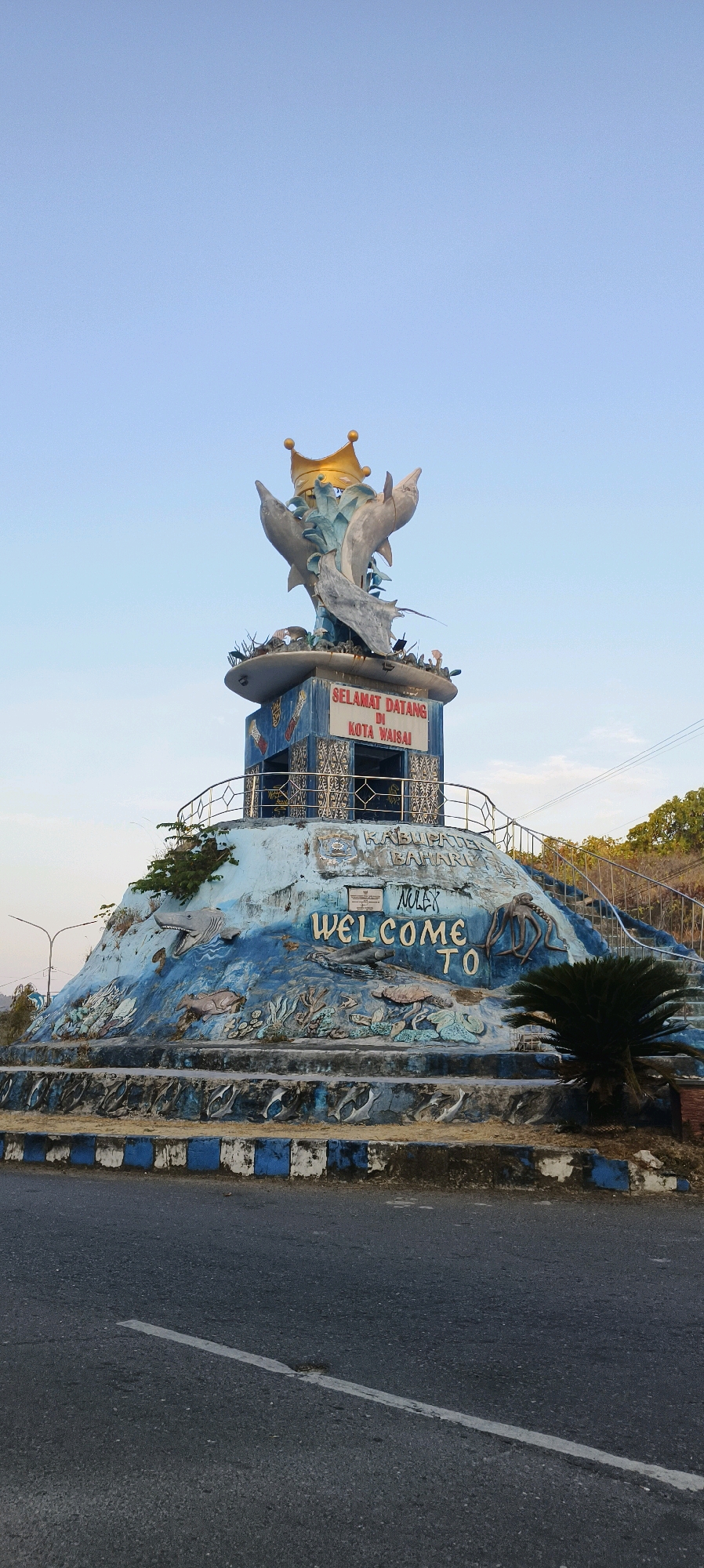 This is a full-frame, eye-level, vertical shot of the Dolphin Monument in Waisai, Indonesia. The monument, which is predominantly blue with white and yellow accents, features a large, ornate gold crown atop two leaping dolphins and a manta ray statue. The monument is situated on a tiered, sculpted base decorated with relief carvings of marine life such as sharks, turtles, and octopuses. A sign on the monument reads "SELAMAT DATANG DI KOTA WAISAI" (Welcome to Waisai City) and "WELCOME TO". The monument is located next to a road, and in the background, there is a clear blue sky with some trees and electrical wires visible. The foreground shows the asphalt road with a white painted line. There are no people or animals visible in the image.