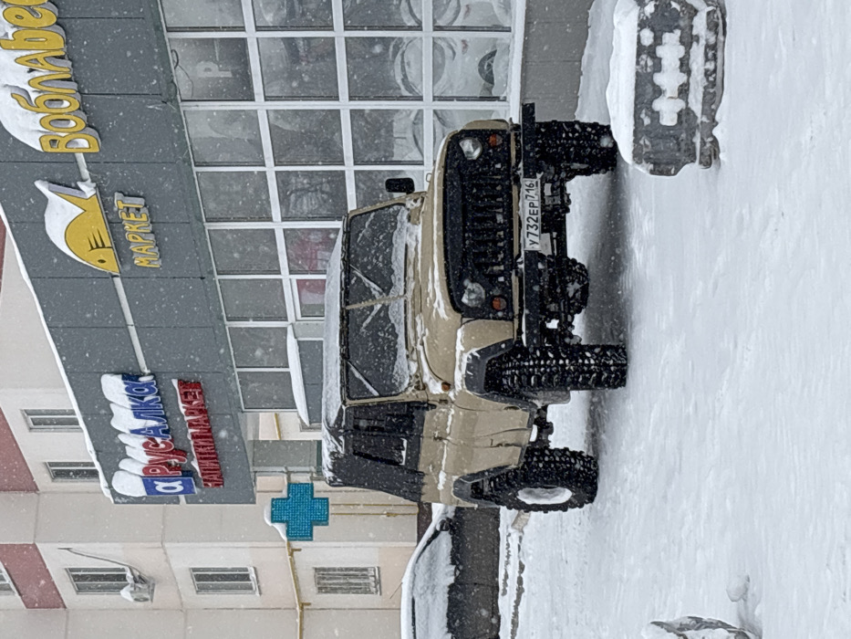 The scene depicts an urban setting in Nizhnekamsk, Republic of Tatarstan, Russia, during active snowfall. A modified tan-colored UAZ-style off-road vehicle, equipped with a black soft top, an aftermarket black grille, and oversized knobby tires, is centrally parked, facing forward. Its windshield, hood, and soft top are dusted with snow. A Russian license plate, Y732EP 716, is affixed to its front bumper. To the left, a dark-colored sedan is partially visible, also covered in snow. The background features a commercial building with a grey paneled facade on the ground floor and large, multi-paned glass windows. Above the windows, Russian signage is visible, including "А Русалка" (A Rusalka) in blue and red lettering with "Напитки Маркет" (Drinks Market) below it. Further right, a yellow fish logo is displayed with "МАРКЕТ" (MARKET) underneath, and partial letters of "Воблабес" (Voblabes) are visible above. A prominent blue illuminated cross sign, indicating a pharmacy, is mounted on the building to the left of the "A Rusalka" sign. The upper story of the building, visible to the left, is light-colored, possibly residential, with multiple windows and an external air conditioning unit. The ground is uniformly covered in a layer of fresh white snow, and snowflakes are visibly falling throughout the frame. In the immediate foreground, to the right, a snow-dusted concrete or stone object sculpted to resemble a razor blade is present. No people are visible.