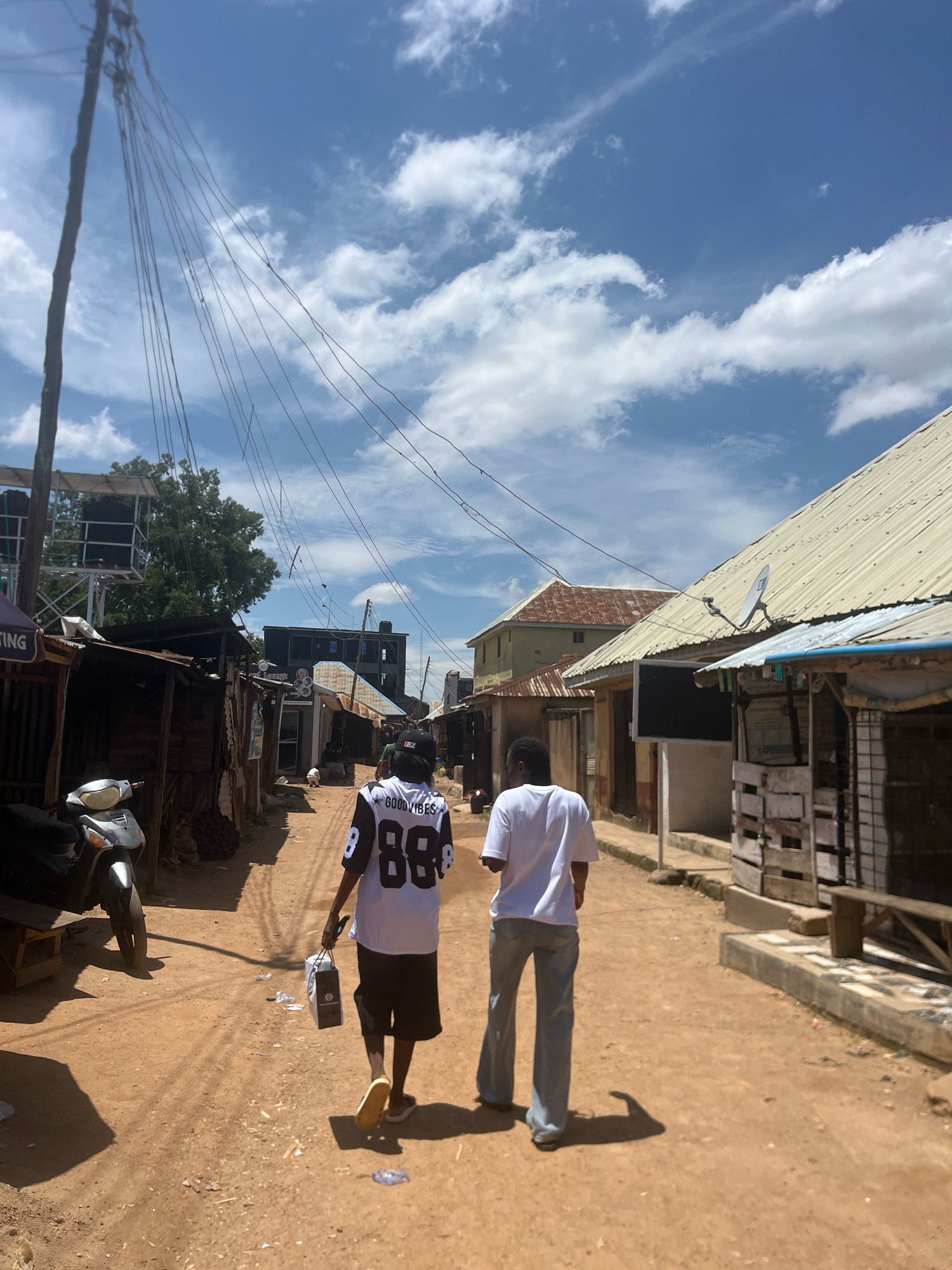 Two individuals walk away from the camera down a dirt road in a densely populated area of Jos, Nigeria, on a sunny day. The road is lined with a mix of structures, including shacks made of corrugated metal and wood, as well as some more substantial buildings. Overhead, a network of power lines crisscrosses the sky, supported by a wooden pole on the left side of the frame. A parked motorcycle is visible on the left, and various items, including plastic bags and debris, are scattered along the road. The sky is blue with scattered white clouds. The person in the foreground wears a white jersey with black sleeves and the number "88" and "GOOD VIBES" printed on the back, paired with dark shorts and light-colored sandals. They are carrying a white bag in their right hand. The second person walks slightly ahead and to the right, wearing a white t-shirt and light-colored flared jeans. Their presence suggests a shared activity or journey down the street. The overall scene depicts a vibrant, lived-in urban environment.