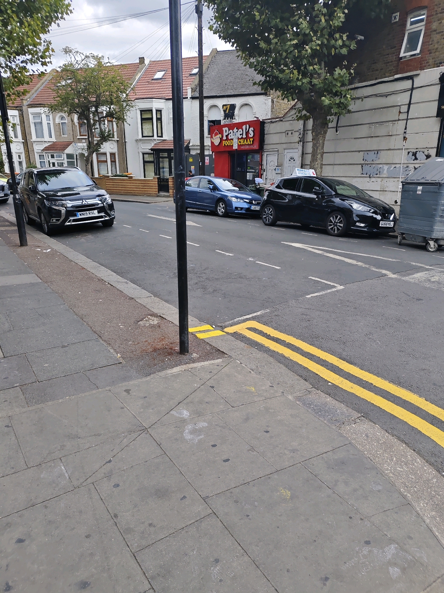 A medium shot captures a street scene in London, United Kingdom, on a cloudy day. The foreground features a grey paved sidewalk with square tiles. Adjacent to the sidewalk is a street with a tarmac surface marked by double yellow lines. Several cars are parked or driving on the street. A black SUV, license plate WN19 RXL, is parked on the left side of the road. Further down the street, a blue hatchback and a black compact car, license plate GU68 NSJ, are visible. A large grey dumpster is situated on the right side of the road. Buildings line the street, including a row of terraced houses and a commercial establishment with a red sign that reads "Patel's Food Chaat." Trees with green foliage are present, and overhead power lines are visible against the sky. The overall scene depicts a typical urban street environment.