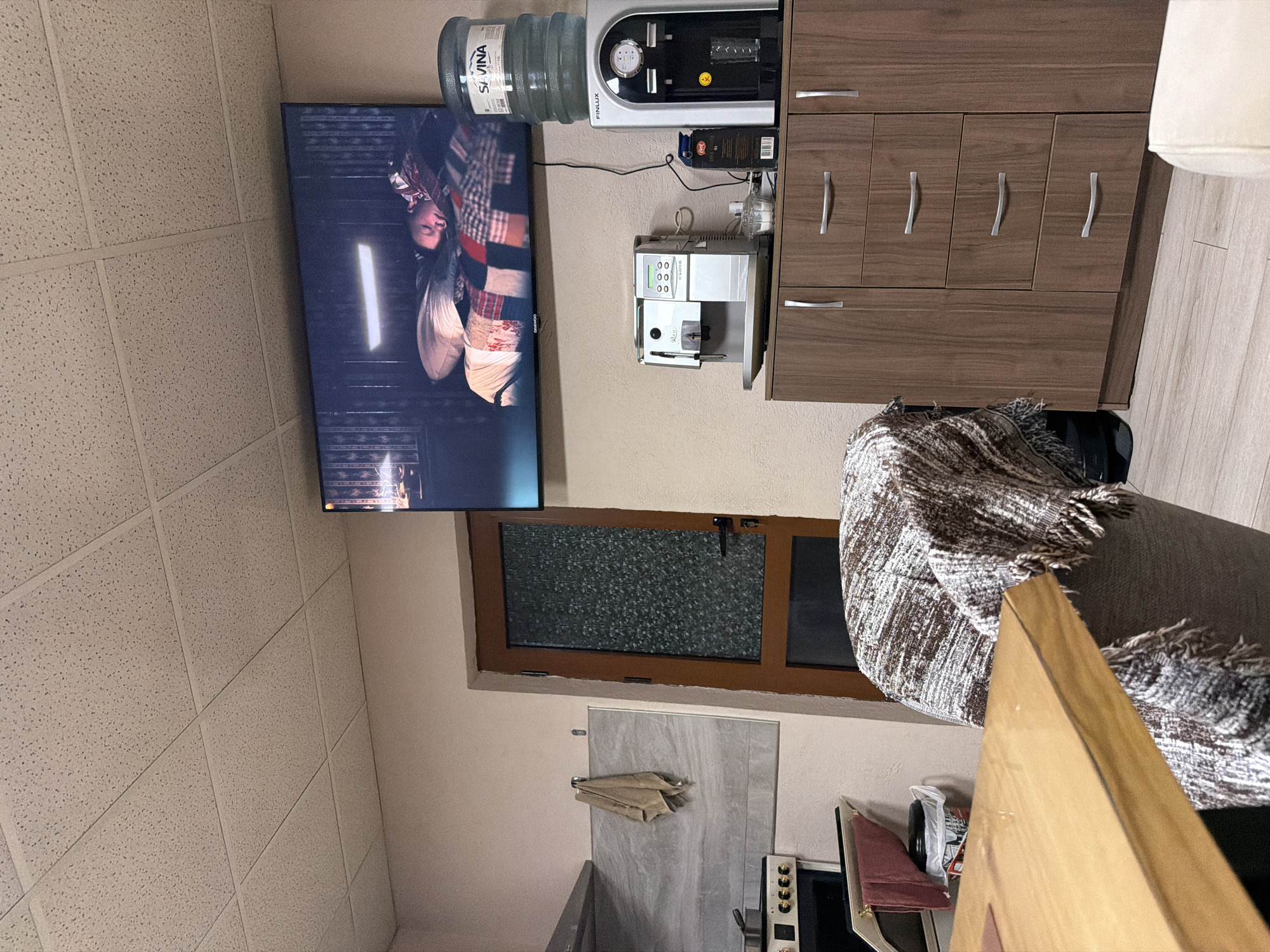 A slightly canted, indoor shot depicts a scene with a blend of residential and utility elements.
The focal point is a wall-mounted flat-screen television displaying imagery that appears to include a person wearing a head covering. Adjacent to the television, and on the same wall, are several appliances: a water dispenser atop a cabinet, a coffee machine, and a cabinet with drawers.
Below the TV and appliances, a window is partially visible with a decorative film covering the glass. The area beneath the window is somewhat obscured by an array of domestic items. These include what appears to be a door or cabinet panel with a towel hanging on it, a kitchen unit, and a table.
To the right, a portion of a blanket-draped armchair or sofa is visible, partially obscuring the view. The floor is not fully visible, but hints of light-colored tiles or wooden planks suggest a clean, contemporary finish. The ceiling has white, square acoustic tiles.
Given the provided location, Panagyurishte, Bulgaria, this appears to be an interior shot of an apartment or home with standard domestic appliances and furniture. There is nothing evident to ascertain an exact address or specific context beyond the general layout and contents of the room.
