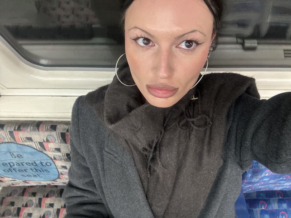 The image displays a rotated close-up portrait of an individual, likely female, situated within a London Underground train carriage in London, United Kingdom. The subject has dark hair, brown eyes with prominent eyeliner and mascara, and full lips with light pink lip color. They are wearing large silver hoop earrings. Apparel consists of a dark grey, possibly wool, overcoat worn over a darker, hooded garment with visible ties or drawstrings at the neck. The individual is directly facing the camera. The background to the left features a patterned fabric seat cover, characteristic of the London Underground, depicting geometric shapes in blue, red, and black against a lighter base. Prominently visible on this seat is a light blue oval sign with white text that reads: "Be prepared to offer this seat," denoting a priority seating area. To the right, a curved train window and a segment of the light-colored interior wall of the carriage are apparent.