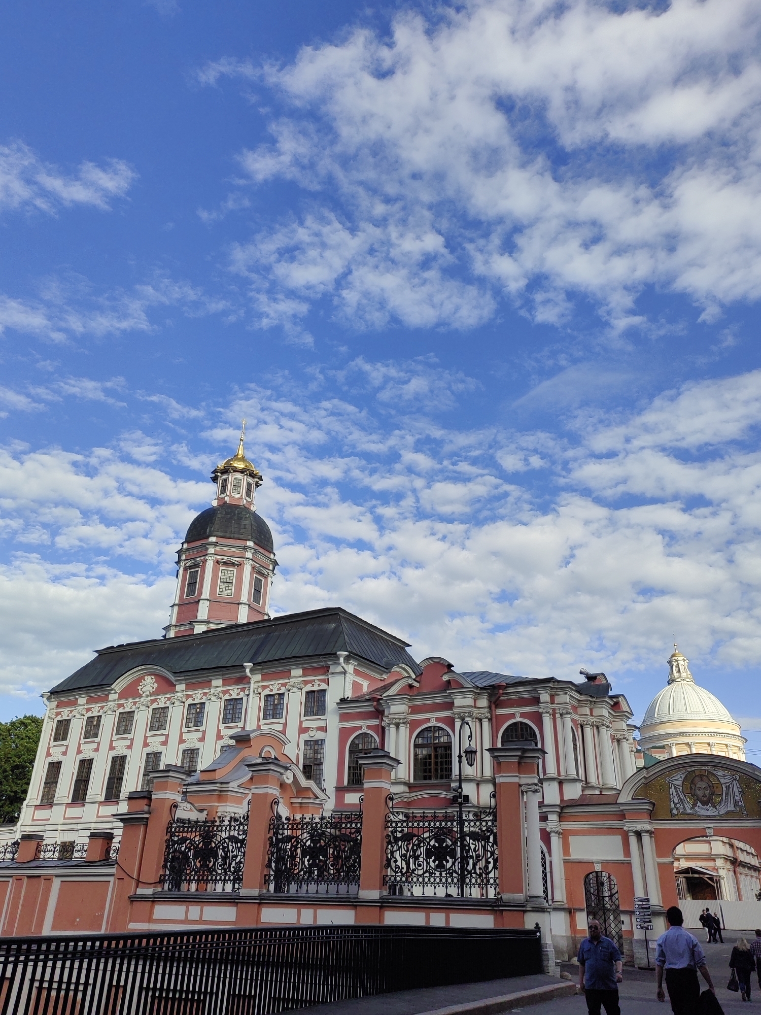 The image captures a view of the Alexander Nevsky Lavra in Saint Petersburg, Russia, on a bright, partly cloudy day. The main subject is the striking architecture of the monastery, characterized by its pink and white facade, ornate details, and multiple domes. A prominent golden-domed bell tower with a black roof stands tall against the blue sky dotted with fluffy white clouds. To the right, a white dome is visible, topped with a golden cross, and below it, an arched gateway adorned with a mosaic of a religious figure.
A decorative pink and white fence with intricate black metalwork separates the monastery from the foreground. In the lower part of the image, a street scene unfolds with several people walking. Two men are in the foreground, one in a blue shirt and the other in a light blue shirt carrying a bag. Other figures are visible further back, suggesting a moderate level of public activity. The time of day appears to be daytime, given the bright sunlight casting shadows. The weather is pleasant, with a clear sky and scattered clouds. The overall impression is one of grandeur and historical significance, set against a picturesque sky. No visible text is present in the image.