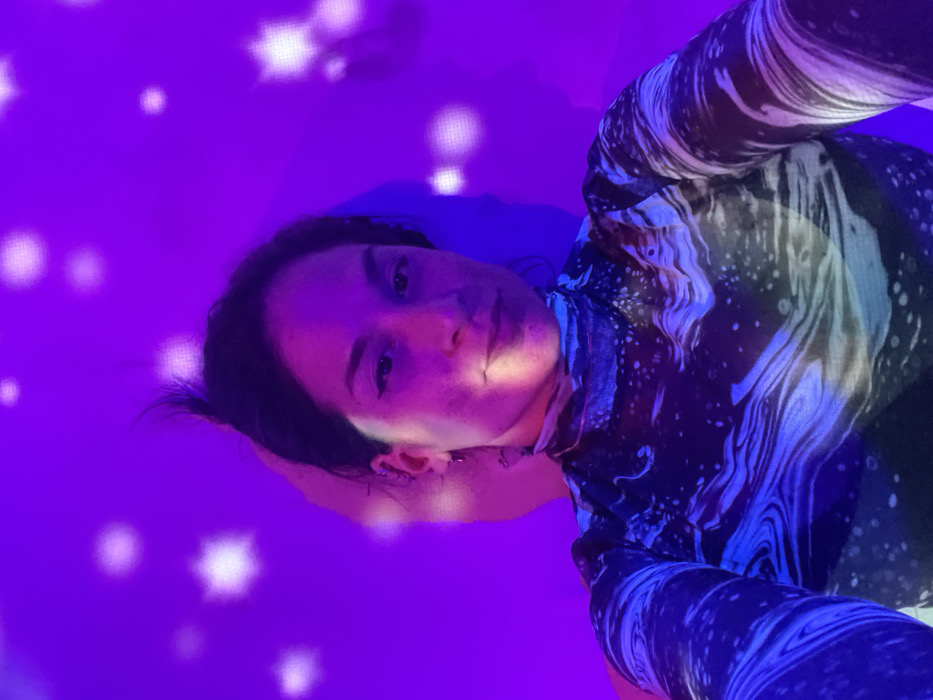 A person wearing a long-sleeved shirt with a marbled or celestial print poses at a tilted angle. The room is bathed in a deep purple light, punctuated by blurry, star-shaped light sources scattered across the background. The light casts strong shadows on the person's face, highlighting one side while leaving the other in shadow.