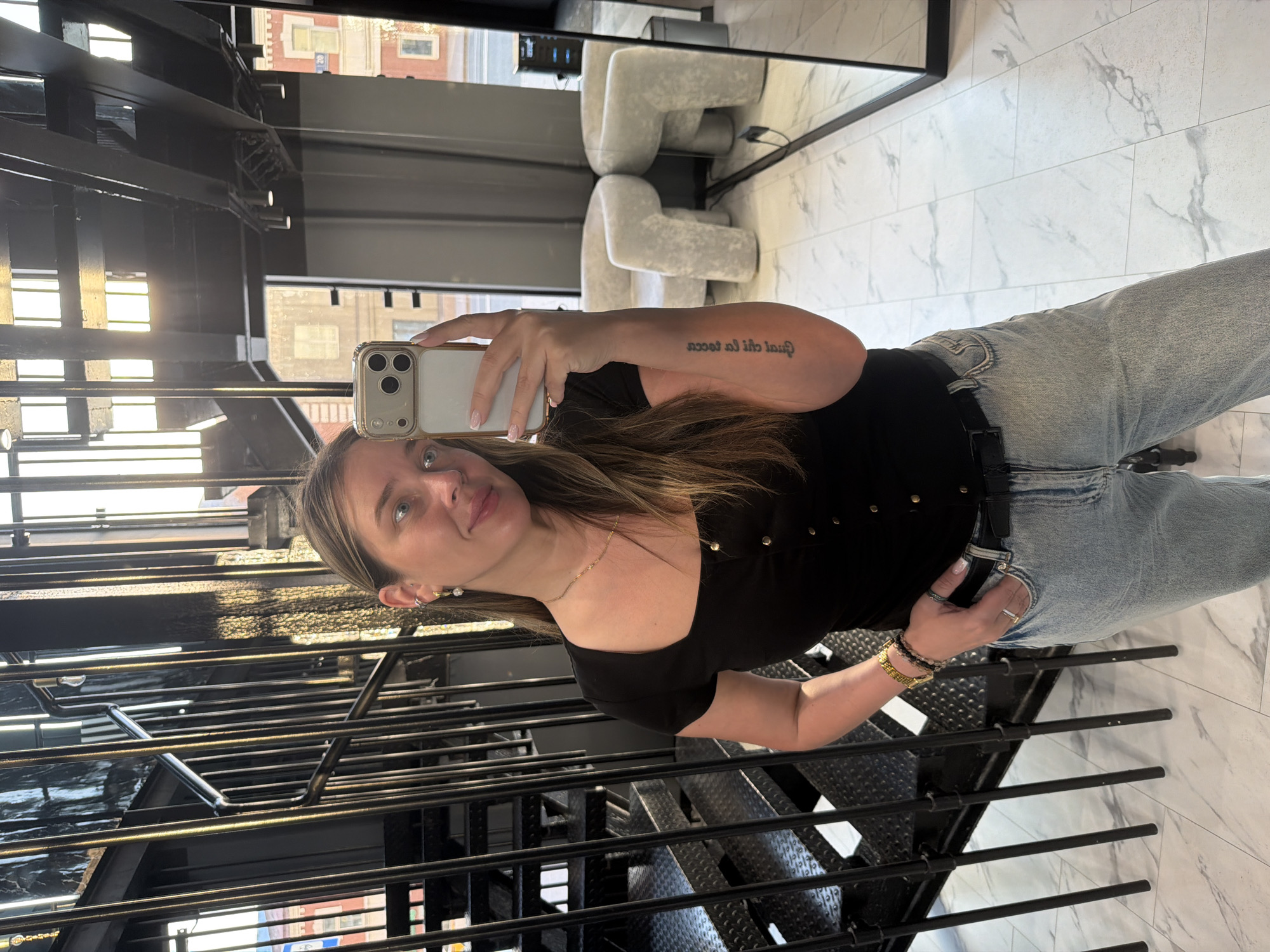 A young adult female is depicted taking a mirror selfie indoors, in what appears to be a modern commercial establishment within the city of Укромновское сельское поселение, Ukraine. She has long, light brown hair with highlights, fair skin, and light-colored eyes, displaying a faint smile. She is wearing a black square-neck top with short sleeves and gold-toned buttons down the front, paired with light-wash denim jeans and a black belt.
Her left ear features multiple piercings, including a hoop earring with a dangling pearl and a silver-colored helix piercing. A delicate gold-colored chain necklace adorns her neck. On her left wrist, she wears a gold-toned watch with a metal strap and several dark beaded bracelets. A silver-colored ring is visible on her right ring finger. Her right hand, holding a light-colored smartphone (identified as an iPhone with a triple-lens camera array) to capture the selfie, shows light-colored nail polish with French tips. A vertical tattoo of non-Latin script is visible on her left forearm, mirrored in the reflection. Her left hand is casually placed in her jeans pocket.
The background features a dark, industrial-style interior. To the left is a prominent black metal spiral staircase with thin balusters and a metal handrail, its steps appearing to have a textured surface. The floor is covered with white marble-patterned tiles. Overhead, a black ceiling with visible ductwork or beams is fitted with recessed lighting. Large windows in the background allow bright natural light to enter, revealing glimpses of external red-brick buildings. To the right, reflected in a large mirror, are two plush, off-white armchairs with distinctive chunky, curved legs. The selfie is captured in a large, framed mirror, with another mirror also visible on the far right reflecting additional parts of the room.