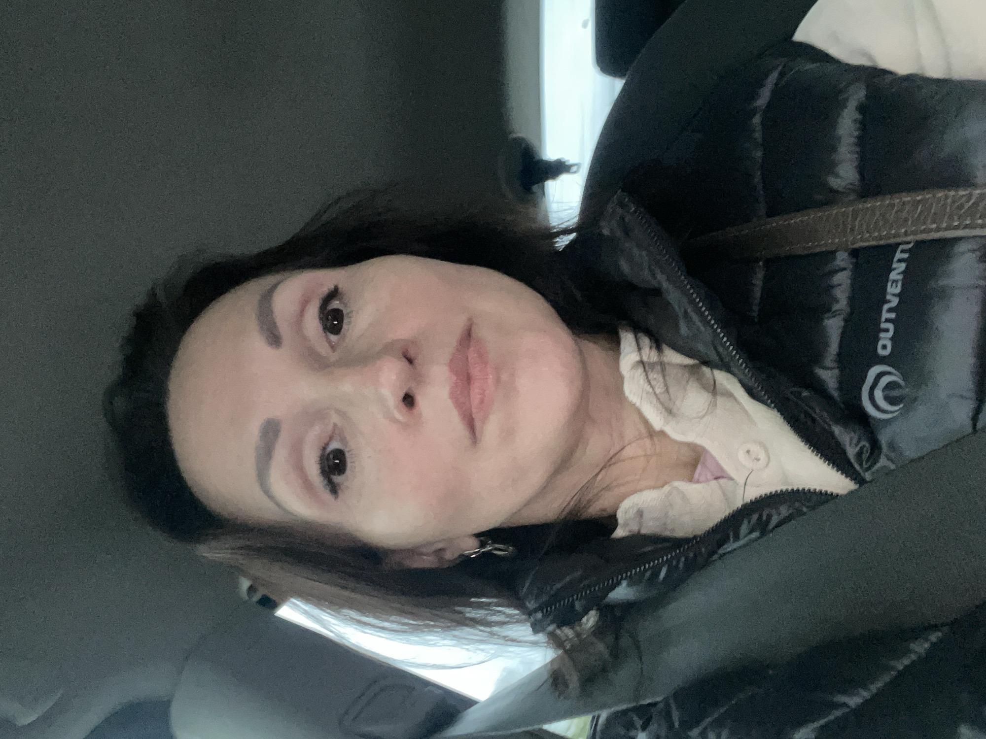 A medium close-up, tilted, selfie shot shows a woman in a vehicle. The interior is gray, suggesting a car. The woman has fair skin, dark hair, and is wearing makeup. She wears a black puffer jacket with a visible "OUTVENT" logo and a partially visible light-colored collared shirt underneath. A seatbelt is fastened across her chest. The location in Varshko, Russia, cannot be determined from this image.