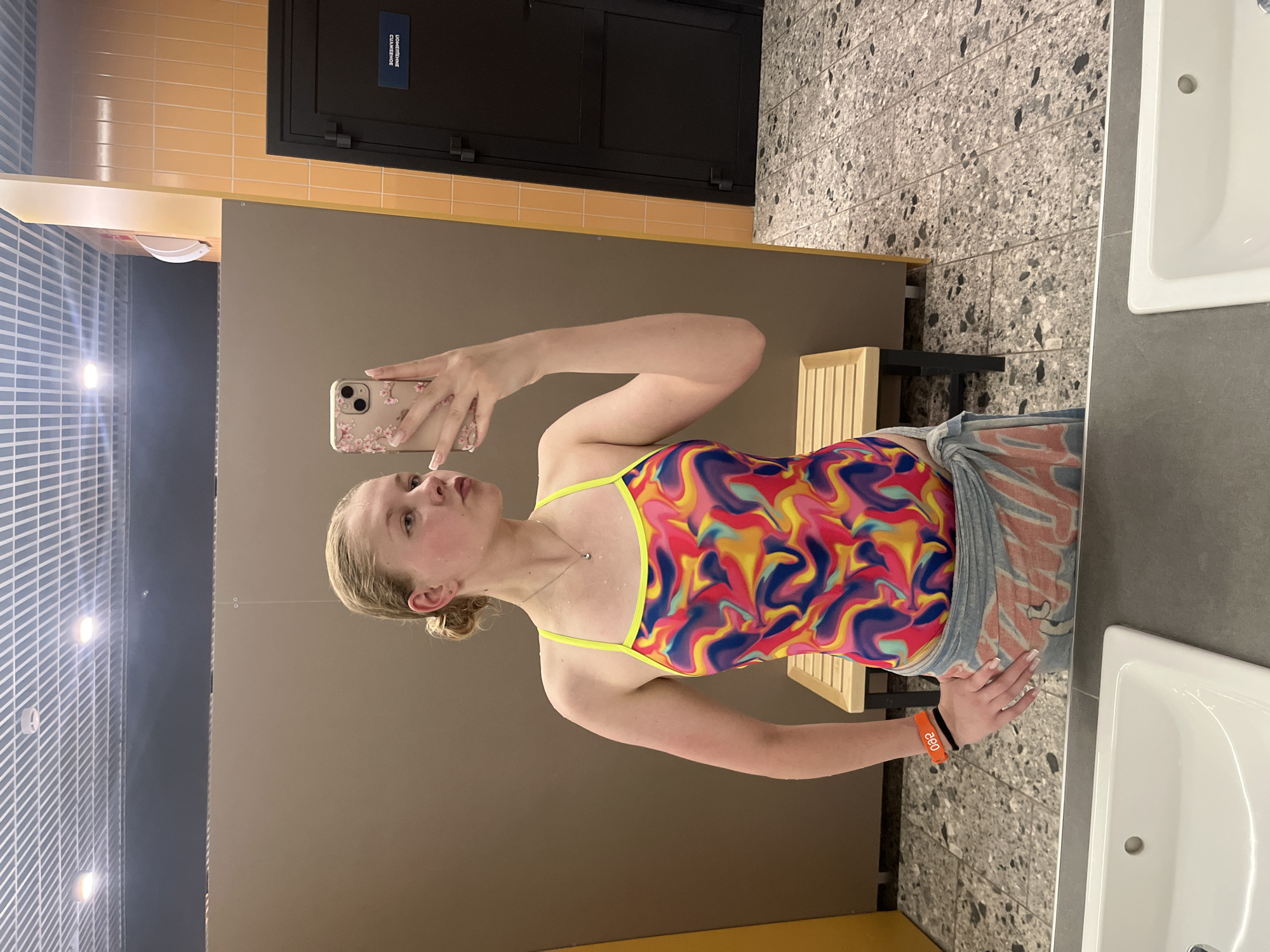 A young woman with blonde hair pulled into a bun stands in a locker room and takes a mirror selfie with her phone. She is wearing a colorful, abstract-patterned swimsuit with neon yellow straps and a light-colored towel wrapped around her waist. She has an orange wristband on her left arm. The locker room has brown walls, orange tiled walls, a black door with a sign in Russian, a tiled floor, and a wooden bench. In the foreground, a white sink is visible. The scene is lit by overhead fluorescent lights.