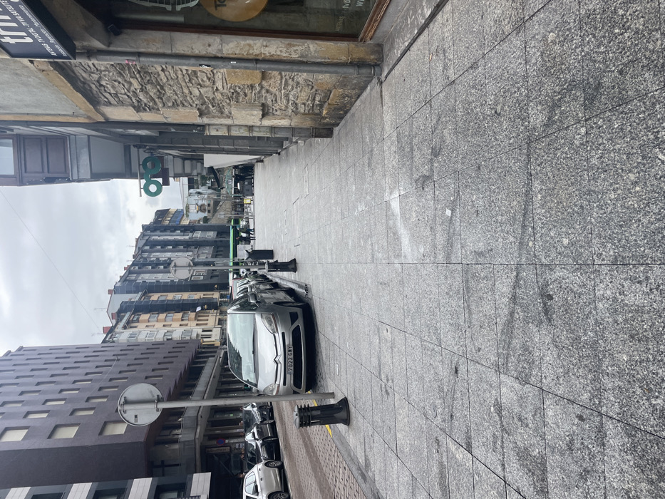 The scene depicts Barrenkale Kalea in Eibar, Spain, under an overcast sky. The foreground features a light grey granite tiled sidewalk exhibiting various dark stains and marks. Along the left side of the street, a silver Citroën C4 Picasso minivan (Spanish license plate 7022 GWV) is parked, oriented away from the camera. Behind it, a series of dark-colored sedans and hatchbacks are also parked. A modern, dark brown multi-story building stands prominently on the left, with older, multi-colored residential buildings extending down the street. Black metal bollards delineate the sidewalk from the roadway, which includes a section of cobblestones transitioning to asphalt further ahead. A circular grey street sign is positioned on a pole adjacent to the bollards on the left. On the right, an older building features a textured stone facade at ground level. A business sign for "TRIA" is visible on this building, advertising "ANTENAS - TV DIGITAL - TV VIA SATELITE." Above the street on the right, a circular sign displaying two interconnected green rings, suggestive of eyeglasses, is mounted. In the distance, near a green city bus, multiple indistinct individuals are visible walking on the sidewalk. No direct interactions between individuals are discernible.