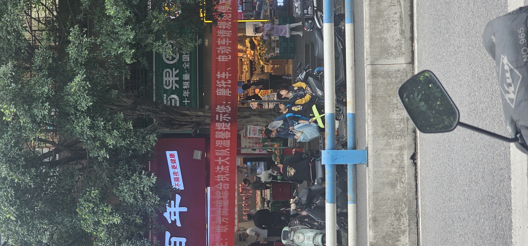 The image depicts a street scene, appearing to be in Shapingba District, China. The view is partially obstructed by a motorcycle mirror and the back of a motorcycle, which are located in the bottom right. The sidewalk has a row of blue and white bollards to separate pedestrians from the road. Behind the bollards, people are walking along the sidewalk and shopping at what appears to be an outdoor market. A red banner with Chinese characters is strung up, and storefronts are visible behind it. A large tree is on the left side of the frame, and the scene is in daylight.