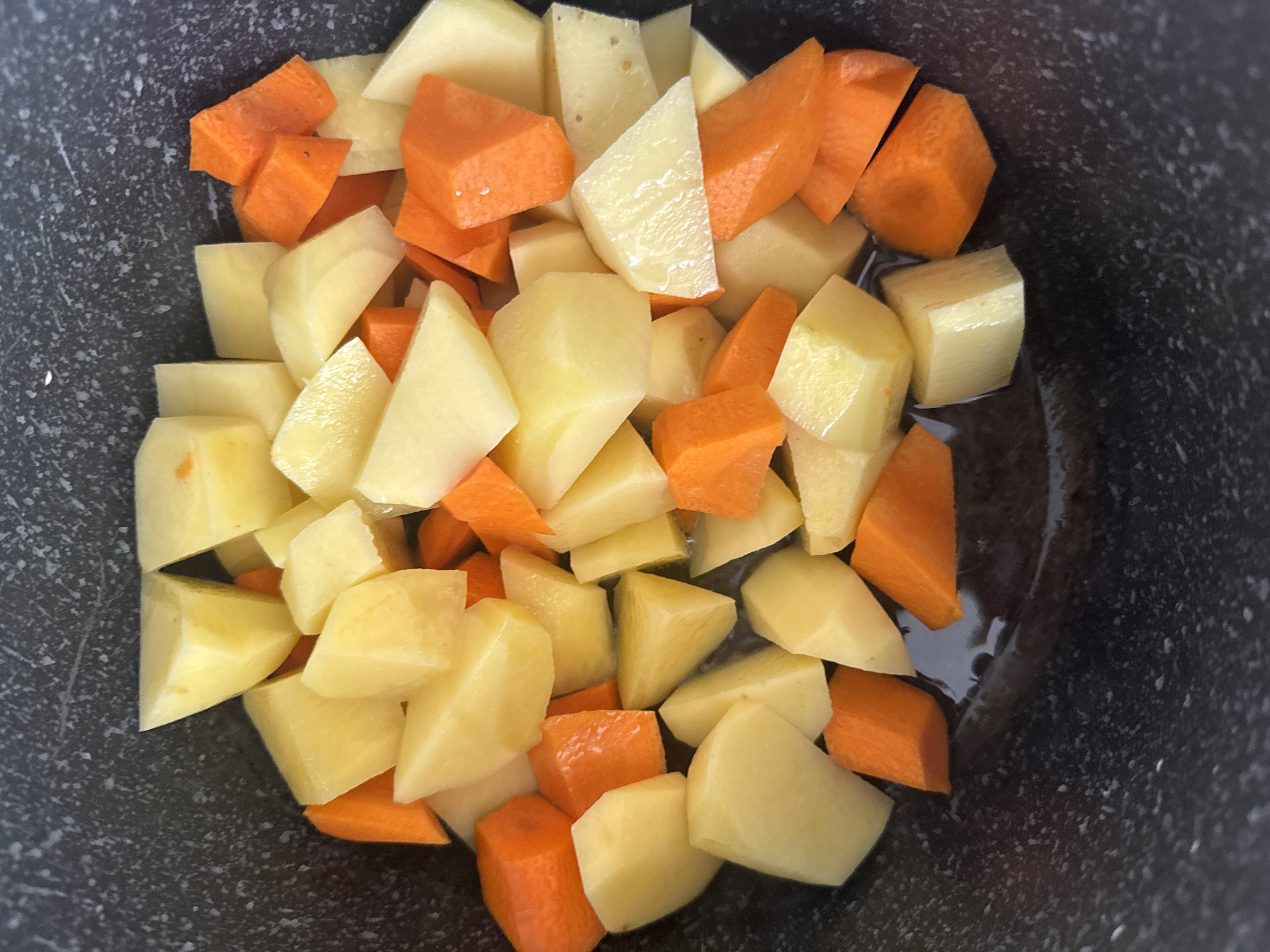 In a close-up, overhead shot, a dark, speckled pot contains chopped potatoes and carrots. The potatoes are cut into roughly cubed and wedge shapes, with their pale yellow flesh visible. The carrots are also cut into similar shapes, displaying their vibrant orange color. The vegetables are mixed together, with some pieces partially submerged in a clear liquid at the bottom of the pot. The lighting is bright, creating highlights on the smooth surfaces of the vegetables. The background is the dark, textured interior of the pot. There are no people or animals present in the image, and no specific location in Bishkek City, Kyrgyzstan can be determined from the visual information.