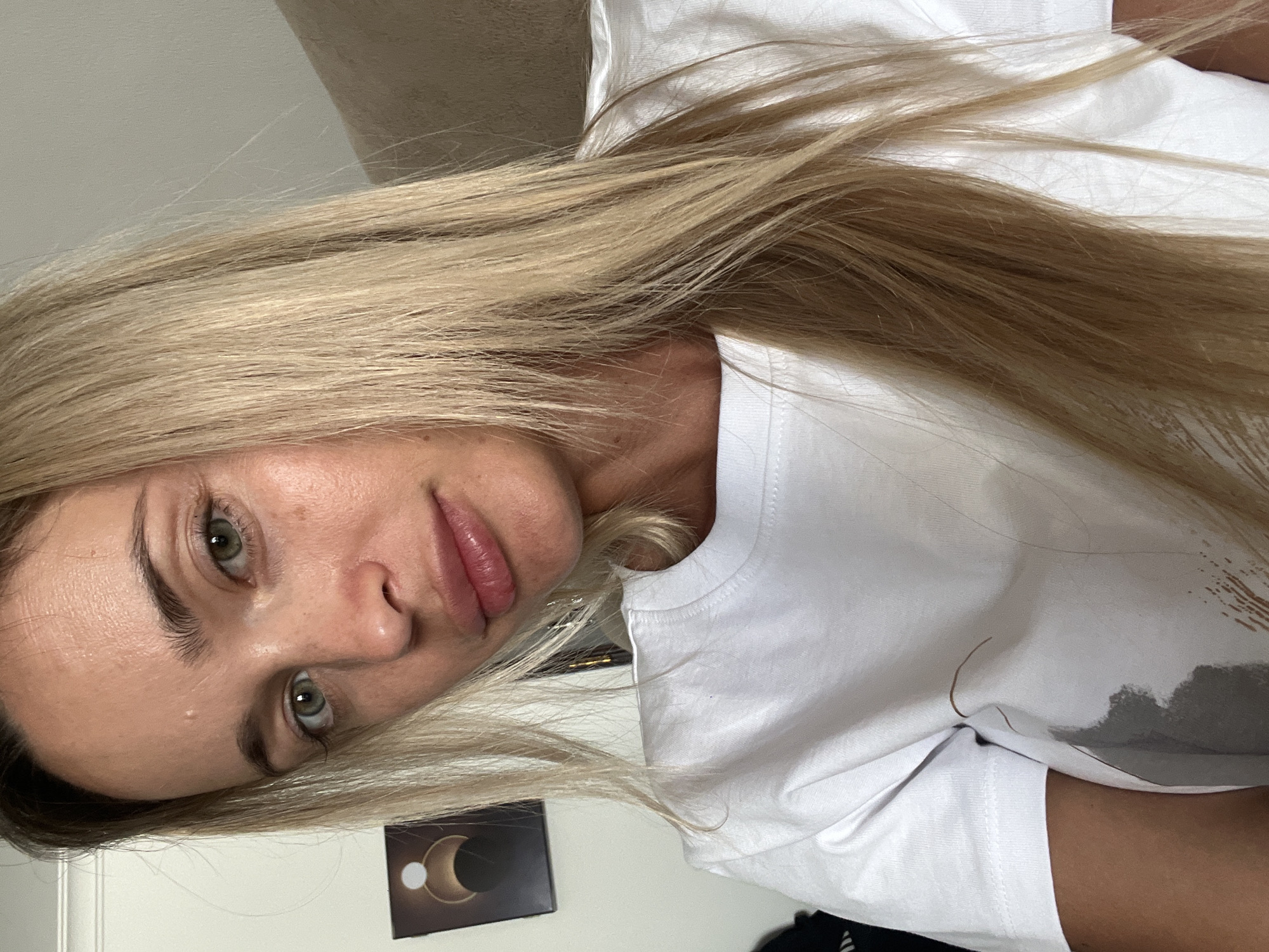 A medium-shot, waist-up photograph shows a blonde woman with fair skin and green eyes. She is looking directly at the camera. She has on a white t-shirt with a gray graphic on the left side. She is wearing lip gloss and has some freckles and blemishes on her face. Her hair is long and straight, falling over her shoulders. In the background, there is a framed picture on the wall and a light-colored sofa. There is no indication of location in Sharm El Sheikh, Egypt.