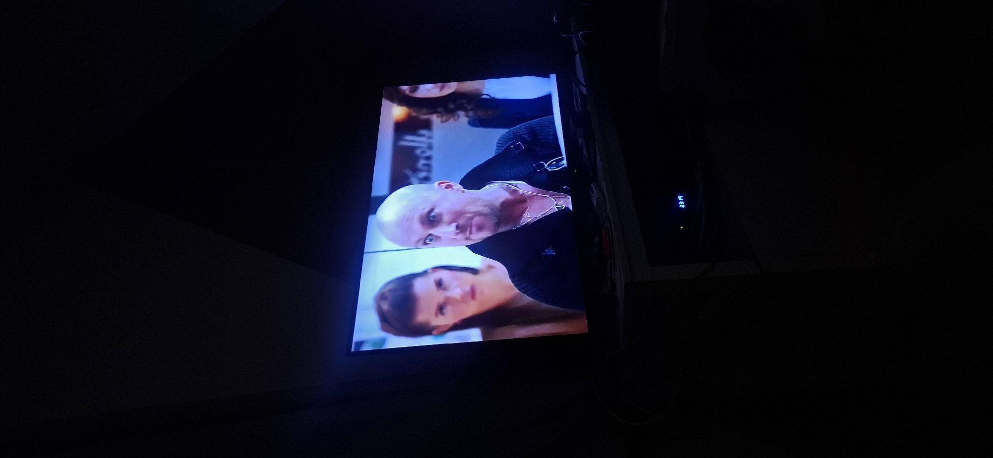 A television screen displays a scene with three individuals. In the foreground, a bald, light-skinned man with a shaved head and a goatee is looking to his right with wide eyes and an open mouth, as if surprised or speaking animatedly. He is wearing a black, textured jacket over a silver chain with two heart-shaped pendants. To his left, a woman with dark hair pulled back and a neutral expression is visible from the chest up. To the man's right, another woman with dark, curly hair is partially visible from the chest up, looking downwards. The background behind the individuals is blurred, but appears to be an indoor setting with some lighting. Below the television, a dark surface is visible, with a digital display showing "23:19". This scene is likely from a residence in Yerocham, Israel.