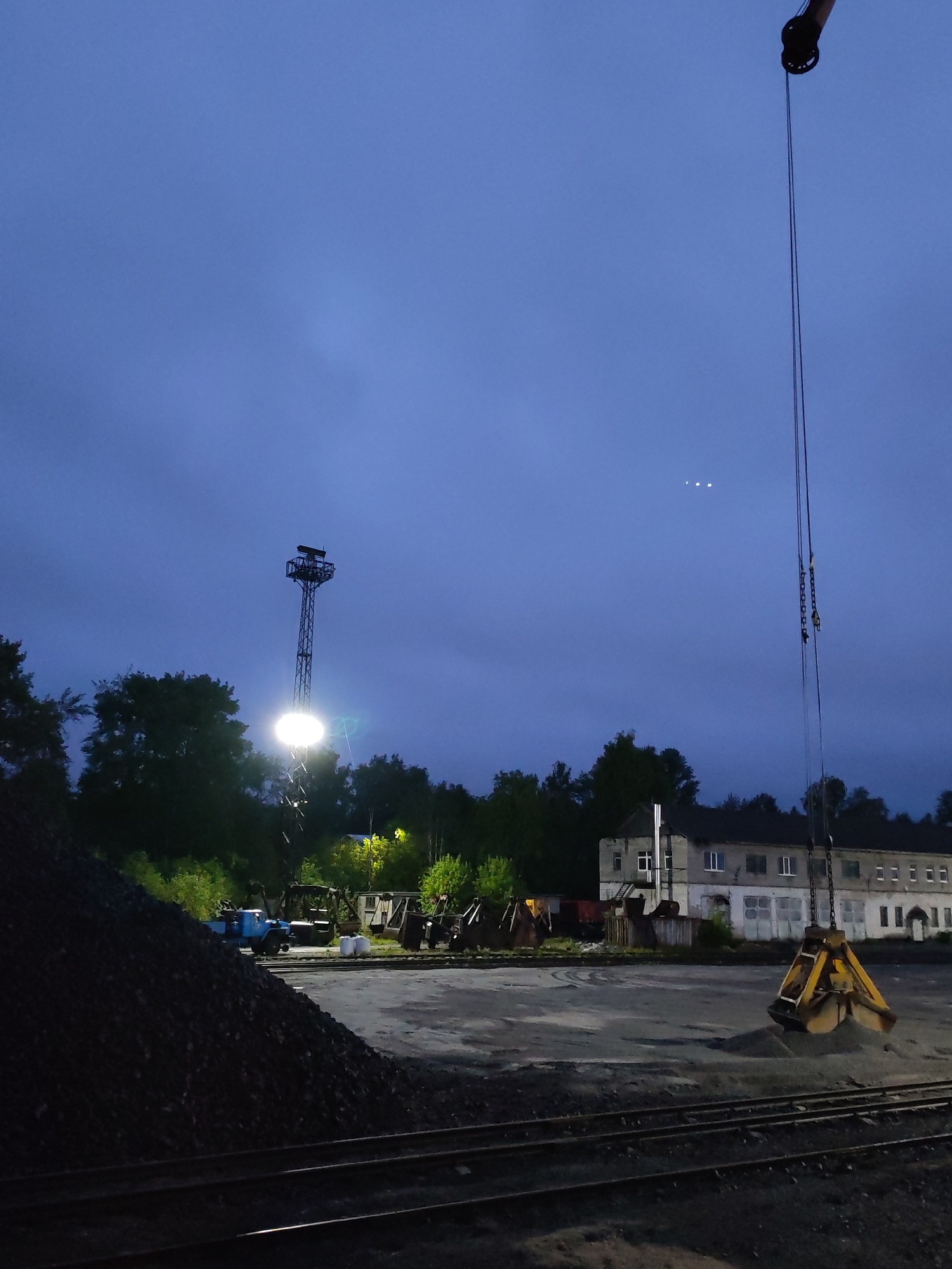 This image depicts an industrial or railway yard scene at dusk or night in Medvezhyegorsk, Russia. The sky is a deep twilight blue, with some visible cloud cover, and three small, bright points of light are discernible in the upper part of the sky.
In the foreground, multiple railway tracks run horizontally across the bottom of the frame. To the left, a large, dark mound of material, likely coal or gravel, dominates the lower-left quarter. In the mid-ground, a tall metal tower with a powerful floodlight stands prominently, casting a bright glow that illuminates the area and creates strong shadows. A blue truck or similar industrial vehicle is faintly visible behind the material pile, and other machinery can be seen in the distance near the floodlight.
On the right side of the image, the boom of a large crane extends downwards from the top, with cables leading to a substantial yellow grab bucket. The bucket is resting on the ground, holding a pile of lighter-colored material, possibly sand or gravel. In the background, an old, multi-story building with multiple windows and a somewhat weathered facade stands behind the crane and grab bucket, with trees surrounding the perimeter of the yard.
The scene suggests an active material handling operation, possibly related to railway transport or storage, occurring under artificial light during the evening or night. No people are visible, and there is no discernible text.