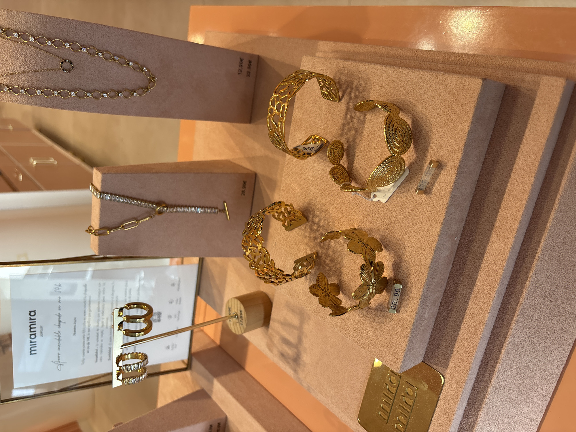 The image displays a close-up, eye-level shot of a jewelry display in a retail setting, potentially in Orihuela, Spain, though this specific location is not definitively confirmed by visual cues within the image. The display is arranged on tiered, light pink, suede-like surfaces.
In the foreground, on the bottom tier, a gold plaque with the embossed "miramira" logo is visible on the left. Adjacent to it, two gold bracelets are displayed. One is a cuff bracelet with a floral design featuring multiple openwork flowers. The other is a wider cuff bracelet with an intricate, woven-like pattern. A small price tag reading "26.99" is attached to the floral bracelet. A small, clear cylindrical object with gold caps, possibly a component for jewelry or a display item, rests to the right of the woven bracelet.
The middle tier features two more gold bracelets. One is a wide cuff bracelet with an open, filigree-like design, and a price tag with "36€" is attached to its interior. The other is a cuff bracelet with an ornate, leaf-like openwork pattern.
On the upper tier, a delicate gold necklace with small, clear rhinestones is displayed on a stand, and a price tag reads "28.99€". To the left, a glass display frame holds a card with the "miramira JEWELRY" logo at the top. Below the logo, text in cursive appears to be in Spanish, describing the jewelry as "Auro increíble chapado en oro 14k". Two pairs of gold hoop earrings are suspended from a thin metal rod in front of this display. The pair on the left is embellished with rectangular clear stones, while the pair on the right is plain.
To the right, a taller, light pink display stand showcases two gold necklaces. The lower necklace is a chain with alternating oval and circular links, interspersed with small clear stones. The upper necklace is shorter and features a circular pendant with dark stones. Price tags are visible on this stand: "12.99€" and "32.99€".
The background shows a blurred view of what appears to be store cabinetry with a pale orange hue, and the lighting suggests an indoor environment. The overall impression is that of a well-organized and attractive retail display of gold-toned jewelry.