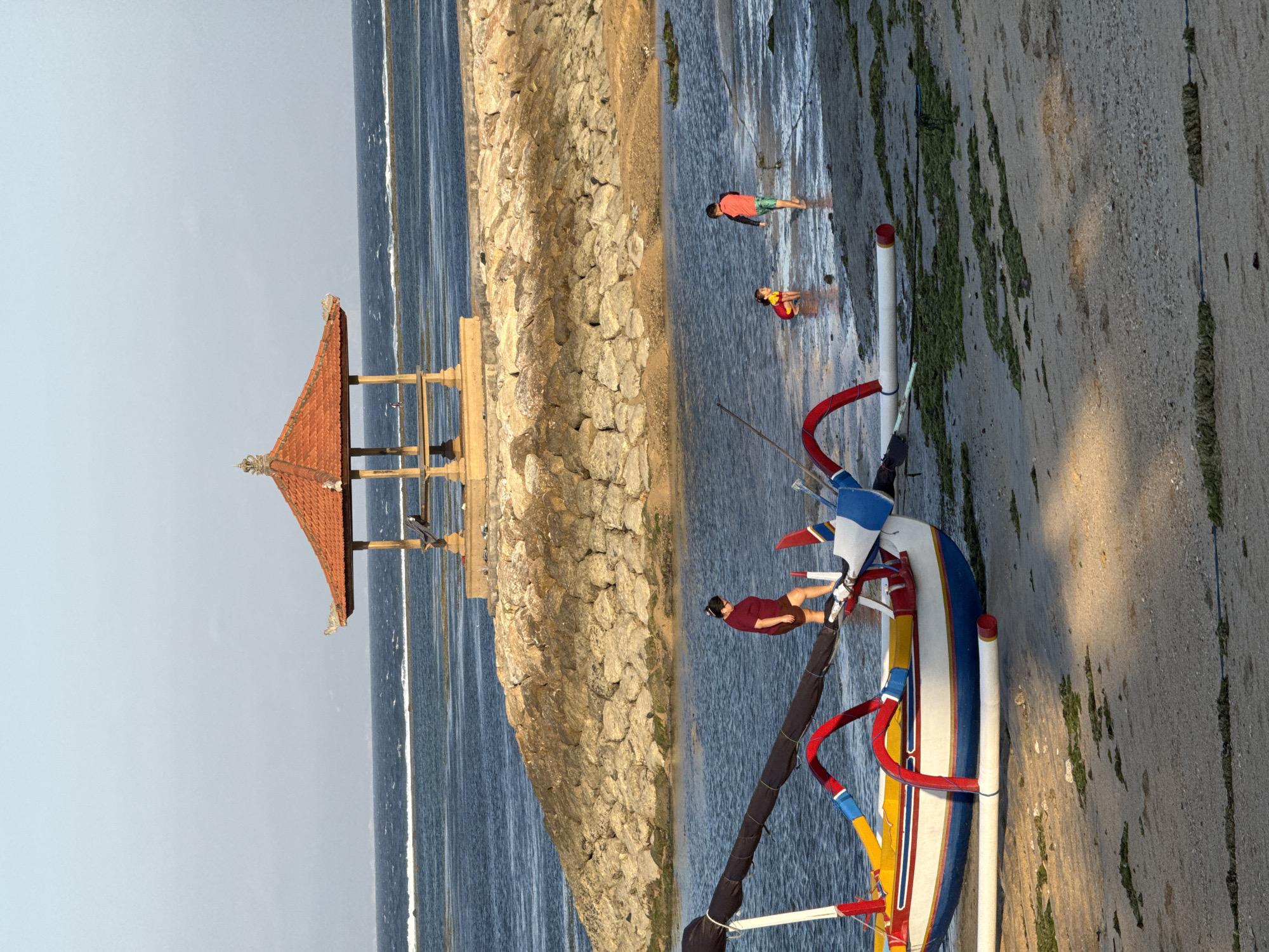 A traditional Balinese Jukung boat, painted with vibrant colors of yellow, red, white, and blue, rests on a sandy beach. The boat has outriggers that extend outwards, supported by curved red and blue structures. In the shallow water near the boat, two children are playing. One child, wearing a red and yellow swimsuit, is kneeling in the water, while another child, in an orange top and green shorts, stands nearby. Further in the background, a person in a maroon shirt and shorts walks on the wet sand. The background features a stone embankment leading to a raised area with a traditional Balinese pavilion. The pavilion has an orange tiled roof and wooden pillars. The sea stretches out behind the pavilion, with gentle waves breaking on a distant reef. The sky is clear and pale blue, indicating it is likely late afternoon or early morning. The scene appears to be located on a beach in Denpasar, Indonesia, a common setting for such traditional boats and architecture.