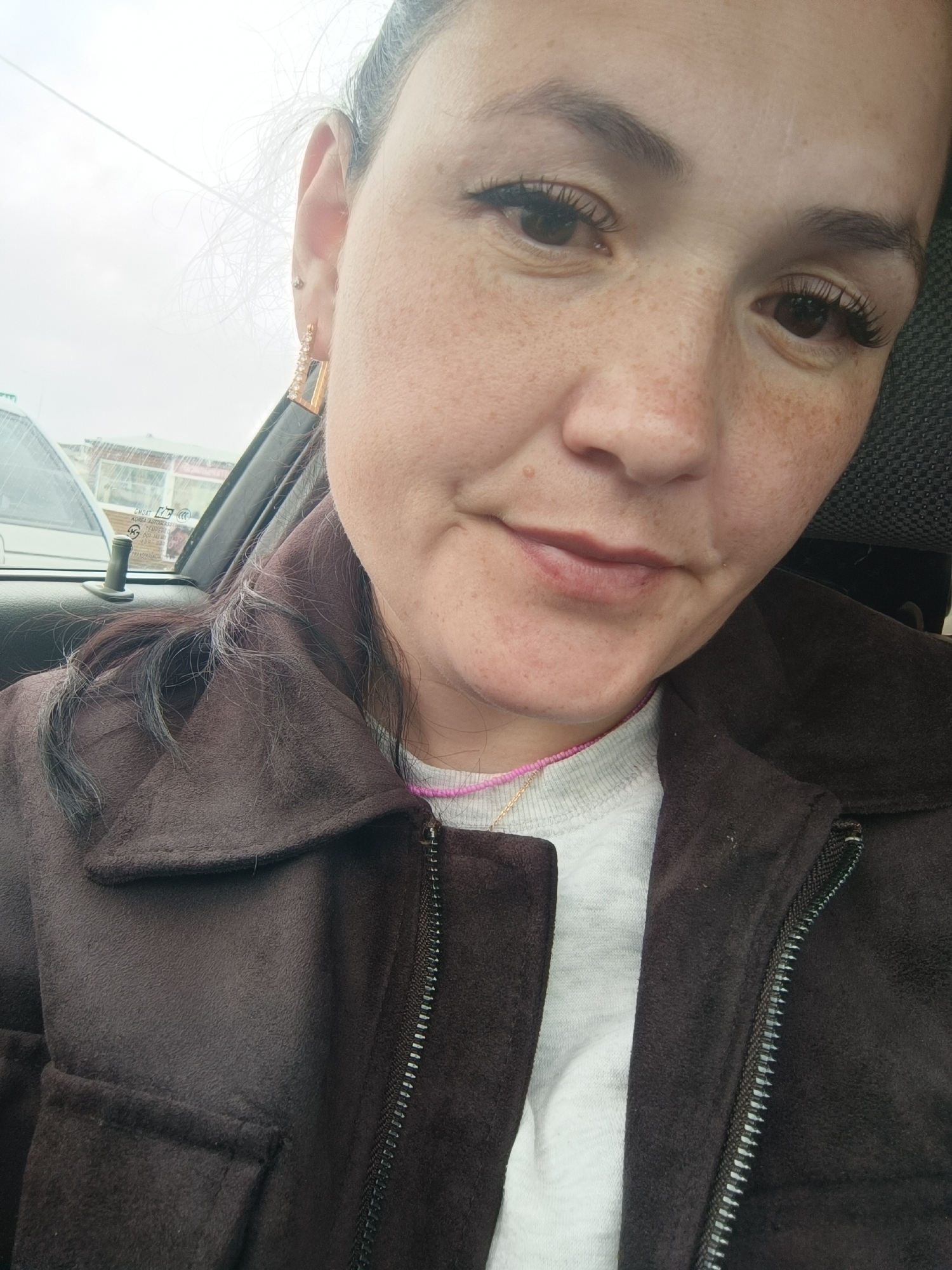 The image is a close-up, selfie-style photograph of a woman in a car. The woman has fair skin with freckles, dark brown hair pulled back, and is wearing eye makeup and a gold earring with clear stones. She is wearing a brown jacket over a light-colored shirt. The background shows a car interior with a car window and a blurred view of buildings and the sky outside. The image appears to have been taken on a cloudy day. There is no specific indication within the image to suggest it was taken in Chaikovsky, Russia.
