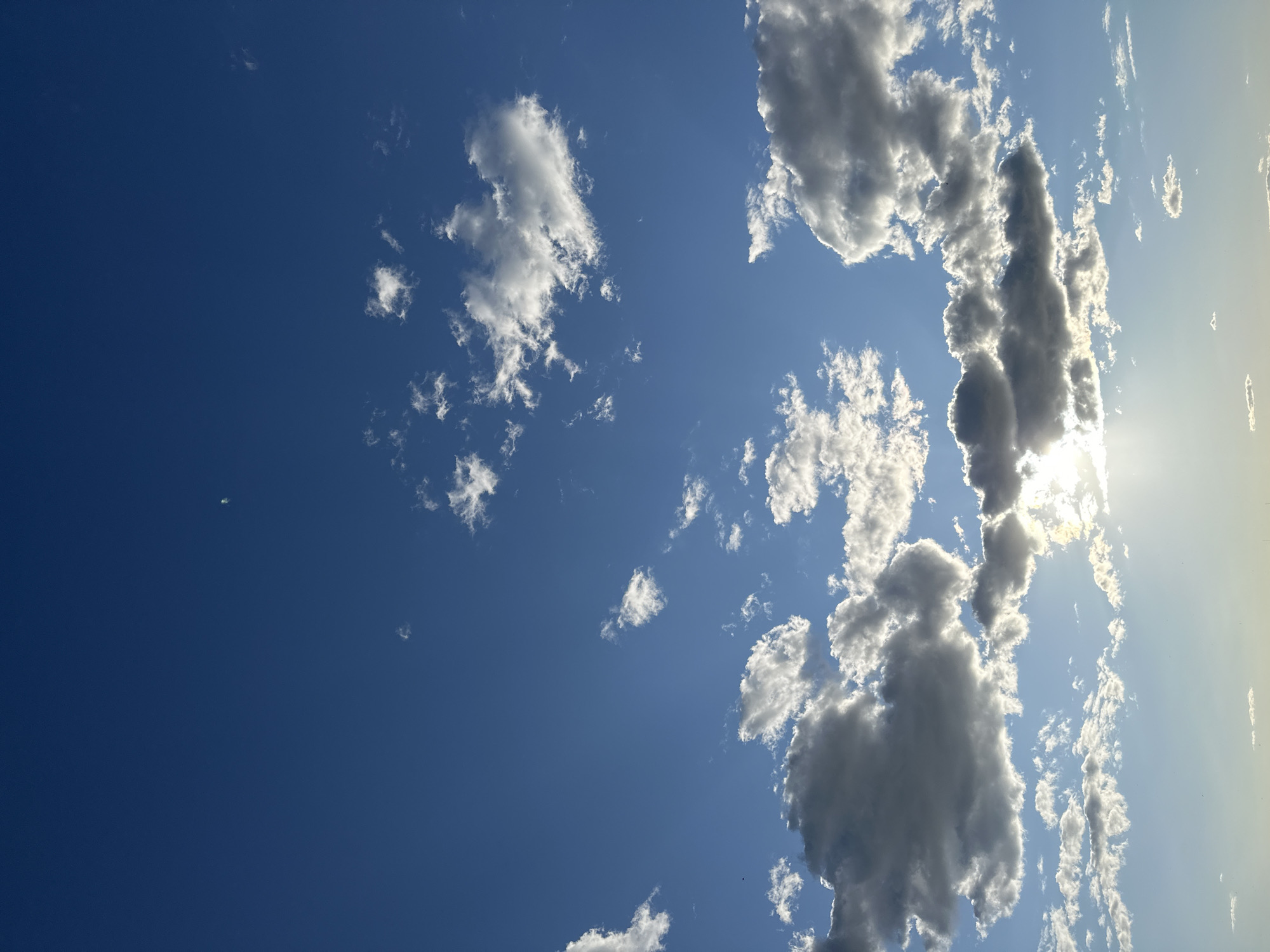 The image shows a daytime view of a blue sky with scattered clouds. A concentration of clouds is present towards the right side of the frame, partially obscuring the sun. The sun's rays are visible through the clouds, creating a bright, diffused light. The left side of the frame is predominantly clear, deep blue sky with only a few sparse clouds. The location is unknown.