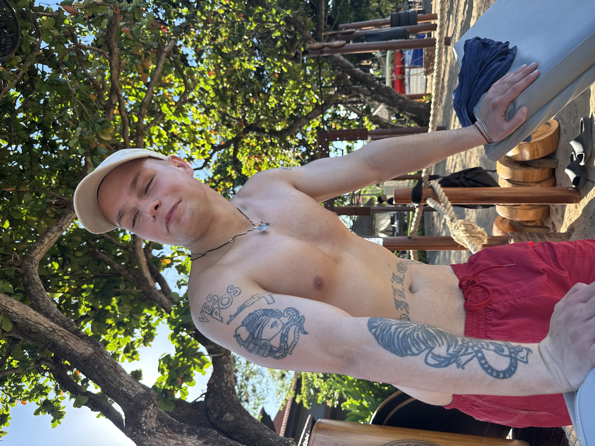 The image shows a shirtless, fair-skinned man with short blonde hair and a light-colored baseball cap. He has several tattoos on his arms and torso. He is wearing red shorts. He appears to be at an outdoor fitness area, leaning against a bench. The area includes wooden workout equipment and appears to be sandy. A tree with green leaves is visible in the background. Based on the visible equipment and foliage, the image location may be in a beach workout area in Kuta Selatan, Indonesia.