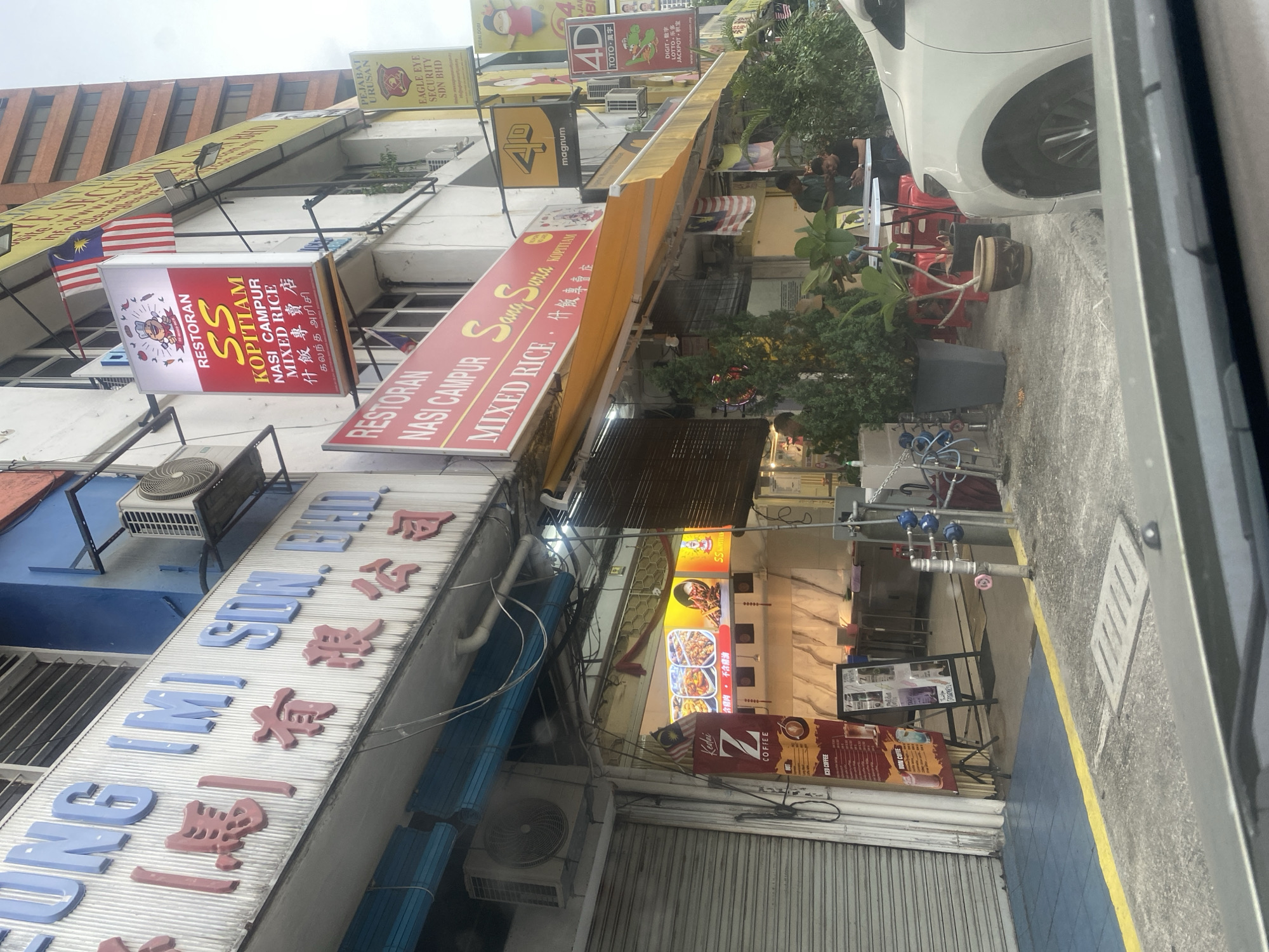 A low-angle exterior shot captures a row of shops in Kuala Lumpur, Malaysia. The foreground is dominated by the hood and windshield of a white car, suggesting the photo was taken from inside a vehicle. The shop fronts, partially obscured by the car, feature various signs and awnings. One prominent sign reads "RESTORAN KOPITIAM SANG SURIA MIXED RICE," with smaller text in Chinese and Tamil below. Another sign displays "RESTORAN SS KOPITIAM NASI CAMPUR MIXED RICE" along with Chinese and Tamil script. Above these, a sign in blue and red lettering appears to be for "UNG (M) SDN. BHD." The buildings themselves are a mix of white, blue, and orange facades.
Several people are visible outside the shops. Two men are seated at a small table with red chairs under a yellow awning, one looking at his phone. Another man stands behind them, interacting with something out of frame. Further to the left, a closed metal roller shutter door is prominent. An air conditioning unit is mounted on the wall above this shutter. A street lamp with a solar panel is attached to the building. Malaysian flags are displayed at different points along the shop fronts. Potted plants and advertising stands are placed near the entrances. The ground is paved with blue tiles and concrete, with a yellow line marking a boundary. In the background, taller buildings with multiple floors are visible.