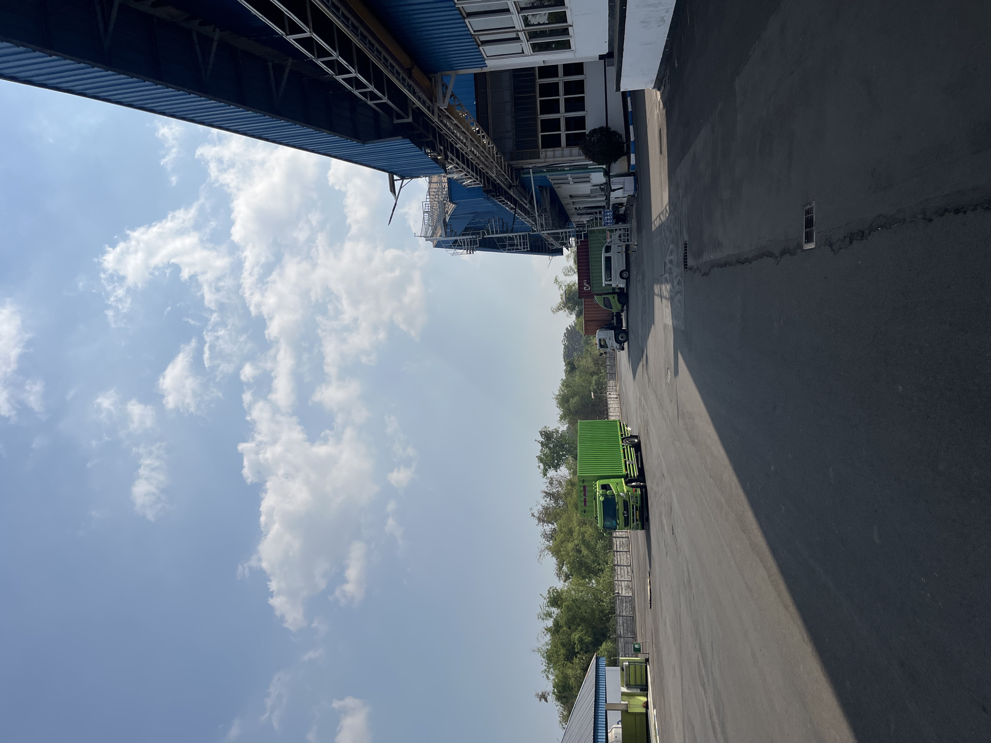 The image depicts an outdoor industrial area, likely within the city of Pelang, Indonesia, under a clear blue sky with scattered white clouds. In the foreground, a dark asphalt road occupies the lower half of the frame, with a prominent shadow stretching across it from the right.
To the left, a light green building with a blue roof is partially visible, adjacent to a concrete fence. Further into the scene, several vehicles are present. A bright green truck with a container is positioned on the asphalt, moving away from the viewer. Behind it, a white truck and a white van are parked or moving slowly.
The dominant feature of the right side of the image is a large, blue industrial building with white trim and numerous windows. A metal walkway or pipe system runs along the side of this building. In the background, a dense line of green trees and foliage forms a natural boundary. The overall impression is one of an active, functional industrial or logistics facility. There are no people visible in the frame.