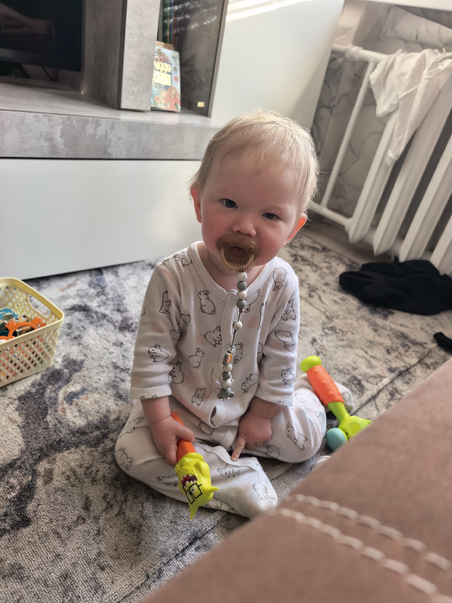 A baby is sitting on a rug in an indoor setting. The baby has blond hair and is wearing a white onesie with a bunny pattern. The baby has a pacifier in their mouth and is holding a yellow and orange toy in their left hand. There is a basket of toys to the left of the baby and a toy next to them on the right. A portion of a pink object is visible in the lower right corner of the image. In the background, there is a white crib and a television stand. The location is not specified, and there is no information to determine if it is in Sterlitamak, Russia.