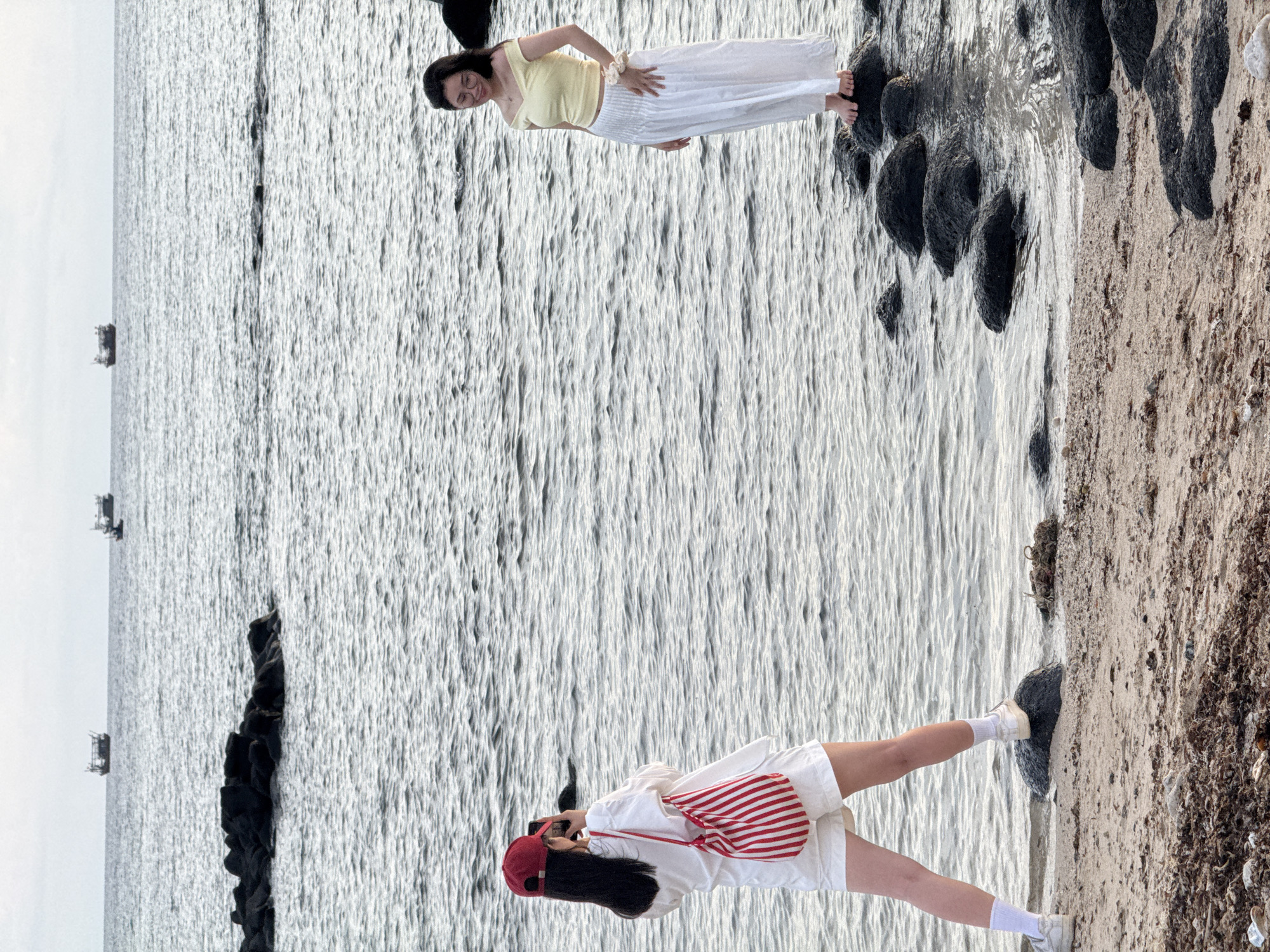 Two women are on a beach in Lý Sơn Special Administrative Region, Vietnam. The woman on the left wears a red baseball cap, a white shirt, white shorts, white socks, and white sneakers. She is holding a phone and taking a picture of the woman on the right. She has a red and white striped bag slung over her shoulder. The woman on the right wears a yellow off-the-shoulder top, a white flowy skirt, and glasses. She is standing on rocks in the water and looking down. The water is calm and a few boats are visible in the distance. The beach is sandy and there are many dark rocks scattered along the shoreline.