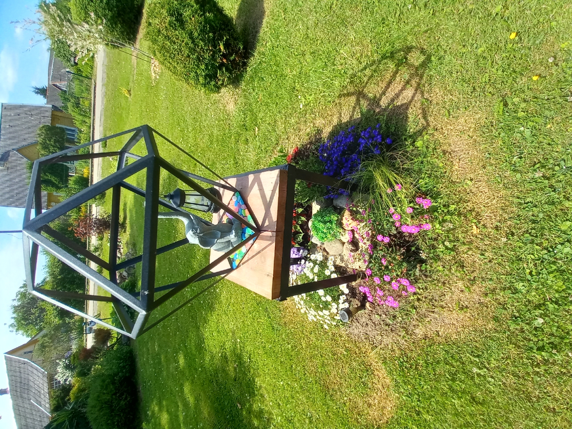 The image depicts a decorative garden feature in Adutiškis, Lithuania. The central element is a wooden table with a black metal frame, topped with a geometric metal structure resembling a geodesic dome. A grey elephant figurine and a black lantern sit on the wooden surface. The base of the feature is surrounded by a rock garden with various small flowers in shades of pink and purple, and some vibrant green foliage. A solar-powered garden light is also visible in the flower bed. The area surrounding the garden feature is a well-maintained lawn. In the background, typical Lithuanian houses with grey roofs and wooden facades are visible, along with trees and a clear blue sky. The overall scene suggests a sunny day in a residential area of Adutiškis.