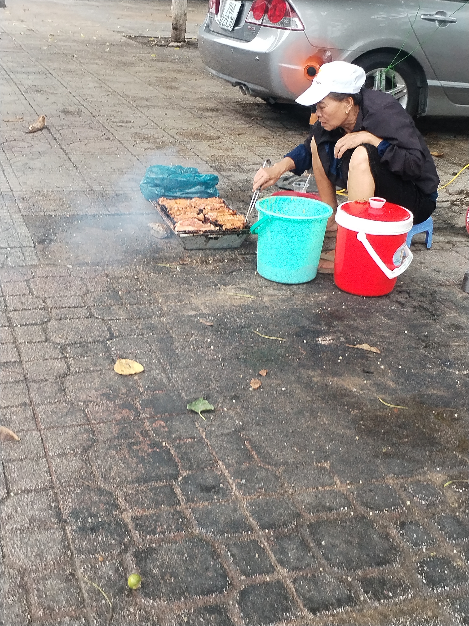 An elderly woman operates a street food stall on a paved sidewalk in Nha Trang, Vietnam, with a silver car parked closely behind her. The woman, wearing a white baseball cap, a dark jacket, and dark shorts, is squatting or kneeling on a small blue plastic stool. She is focused on grilling pieces of what appears to be marinated meat on a rectangular charcoal grill, using metal tongs to turn the food. Wisps of smoke rise from the grill.
Beside her are two plastic containers: a bright turquoise bucket to her left and a red insulated container with a white lid and handle to her right. A teal plastic bag is also visible near the grill. The paved ground beneath them is discolored and shows signs of wear and grime.
The license plate of the car behind her is clearly visible as "79A 229.30," with "79A" indicating Khánh Hòa province, where Nha Trang is located. The lighting is diffused, suggesting it is daytime under an overcast sky or possibly late afternoon. The scene depicts a common sight of street food vending in Vietnam. No strong emotions are discernible on the woman's face beyond concentration.