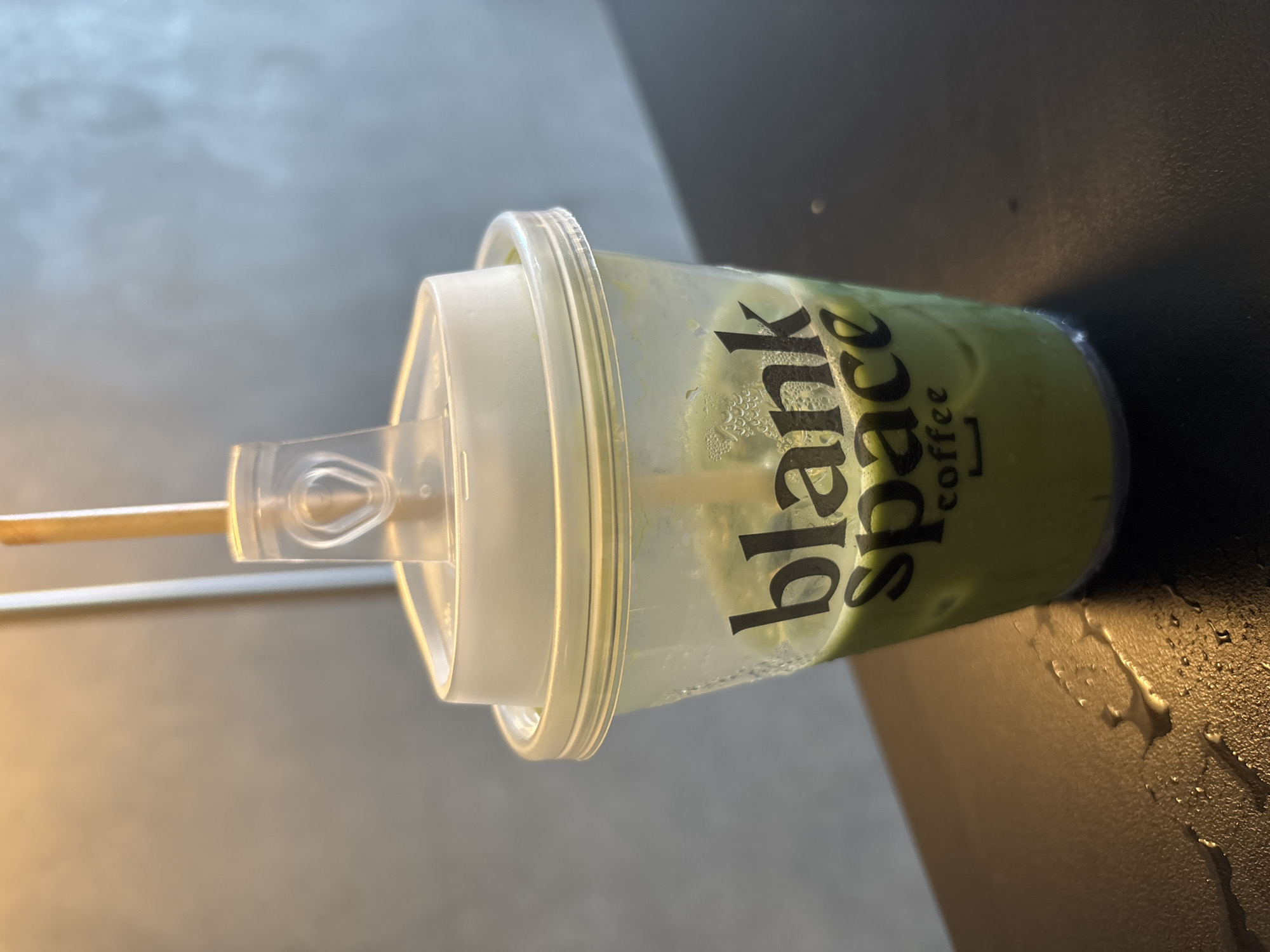 An iced matcha latte in a clear plastic cup with a white lid and a light brown straw sits on a dark, wet surface. The cup has "blank space coffee" printed in black text on it. The background is a grey, textured wall. The scene is indoors, likely in a cafe or coffee shop. The overall impression is one of a refreshing drink on a potentially warm day. It is not possible to determine the specific location within Davao City from the image alone.