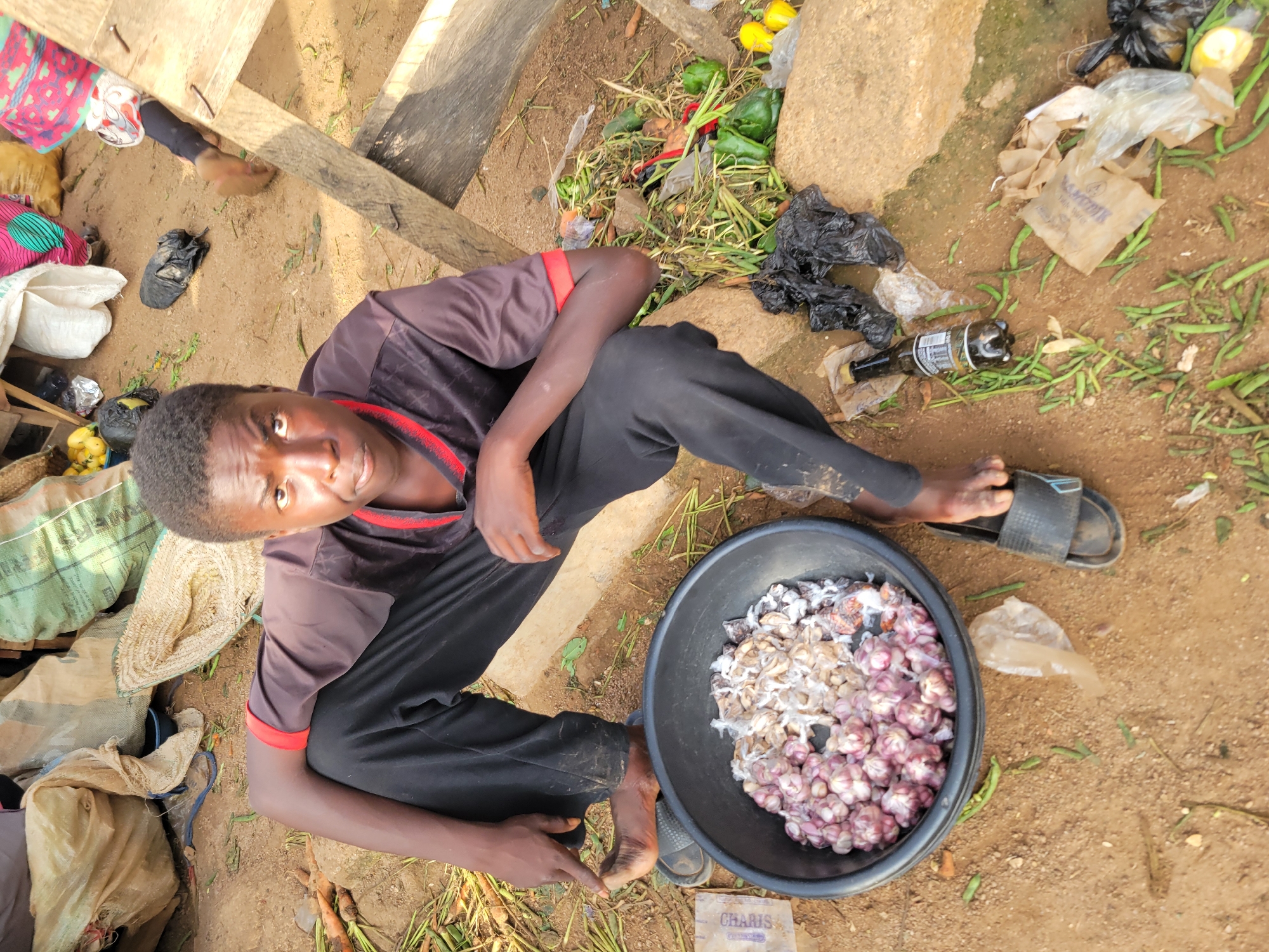 A young person wearing a black t-shirt with red trim and black pants is sitting on the ground next to a large black bowl filled with garlic. The person is barefoot, wearing black flip-flops. The bowl of garlic is in front of them, and to their right, there is a collection of green vegetables, including bell peppers and what appear to be green beans. Several plastic bags, some containing produce and others empty, are scattered around the scene, along with some loose vegetables on the ground. A bottle of dark liquid is also visible on the ground near the vegetables. The ground is dirt, and in the background, there are large sacks, possibly containing grains or other goods, and wooden structures. The overall environment appears to be an outdoor market or roadside stall. The lighting suggests it is daytime.