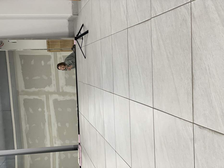 The image depicts an indoor scene in Budva, Montenegro, characterized by a light-colored tiled floor and a large mirror occupying the background. A young adult female is visible in the mirror's reflection, positioned to the right. She has light brown hair, is wearing a light grey long-sleeved shirt, and is holding a dark smartphone up, appearing to take a selfie. Her lips are slightly pursed. The mirror reflects an unfinished wall behind her, showing light grey drywall with visible seams and patches. To the left of the woman in the reflection, pink rectangular foam blocks are visible on the floor against another reflective surface. To the right of the woman in the reflection, a light brown wooden slatted structure is partially visible. A black tripod with three legs stands on the tiled floor in the midground, between the viewer and the mirror. The immediate wall to the right of the mirror is white.