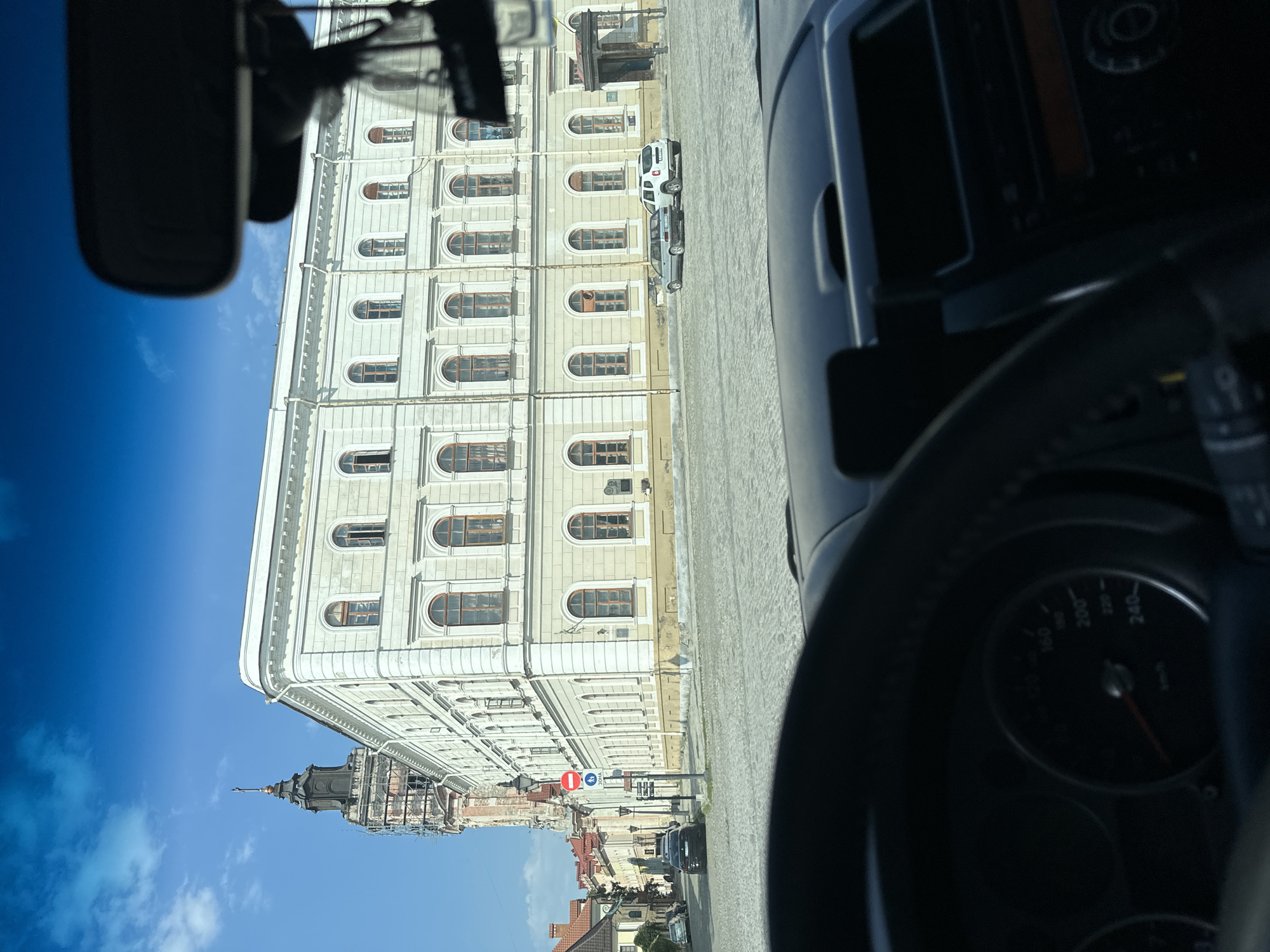 The image, taken from inside a vehicle, shows a view of the historic city center of Kamianets-Podilskyi, Ukraine. The foreground is dominated by the steering wheel and dashboard of the car, with a speedometer visible showing a reading around 30 km/h. The windshield reveals a cobblestone street and a large, light-colored building with arched windows and classical architectural details. To the left, a tall, dark-colored tower with a spire, possibly part of a church or castle, is visible, with scaffolding around its upper section, suggesting ongoing construction or restoration. Several cars are parked on the street and in the background, and streetlights are present. The sky is blue with scattered white clouds. The overall scene depicts a sunny day in a European city square.