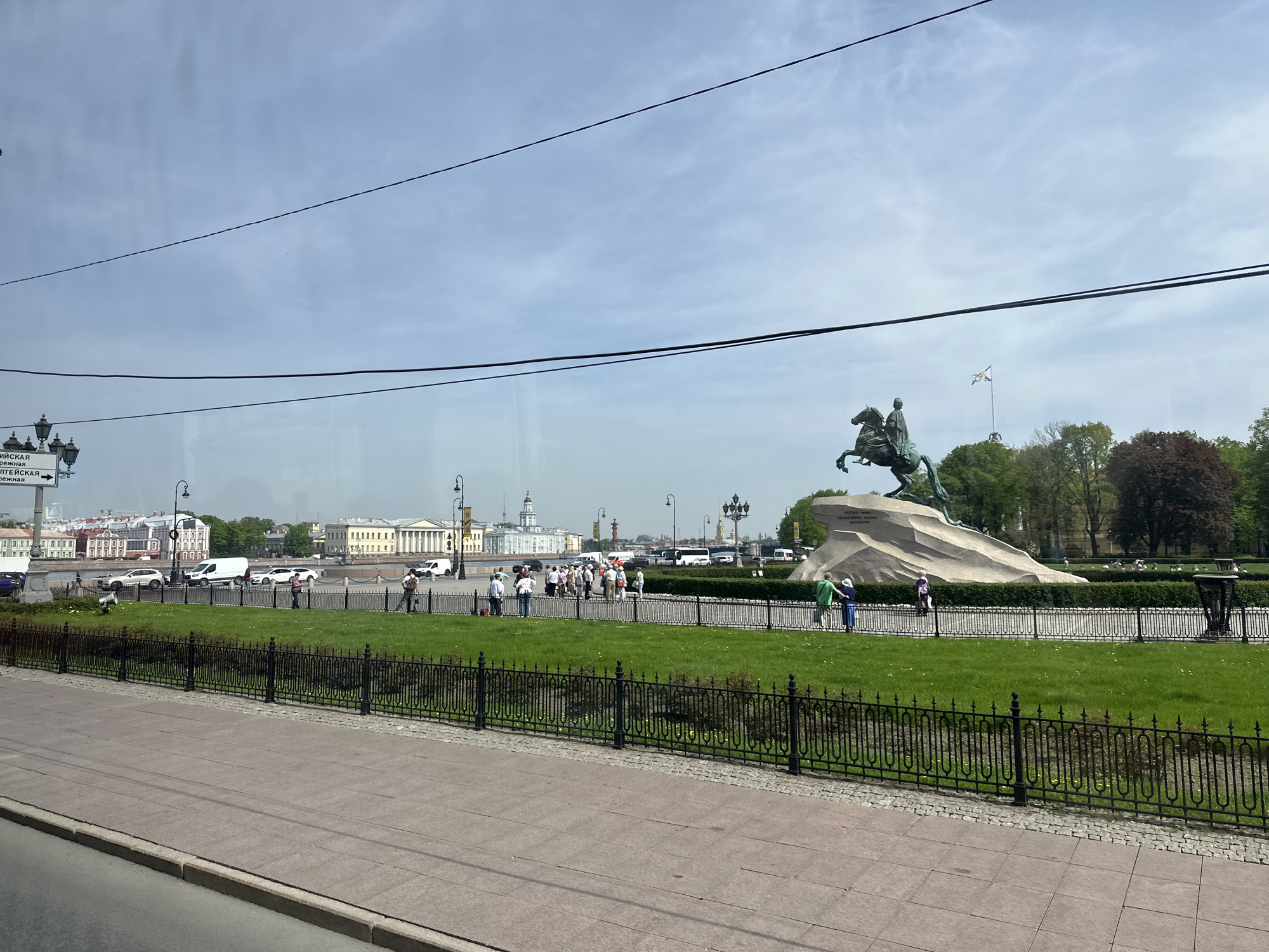 The image shows a view of the Bronze Horseman statue in Saint Petersburg, Russia. The statue depicts Peter the Great on horseback, a prominent monument in the city. The image is taken from a slightly elevated position, possibly from a moving vehicle. A group of people are walking along a pathway near the statue. The Neva River and some buildings are visible in the background. Overhead electrical wires are visible in the sky. The weather appears to be fair with some clouds. A fence separates the sidewalk from the grassy area around the statue.