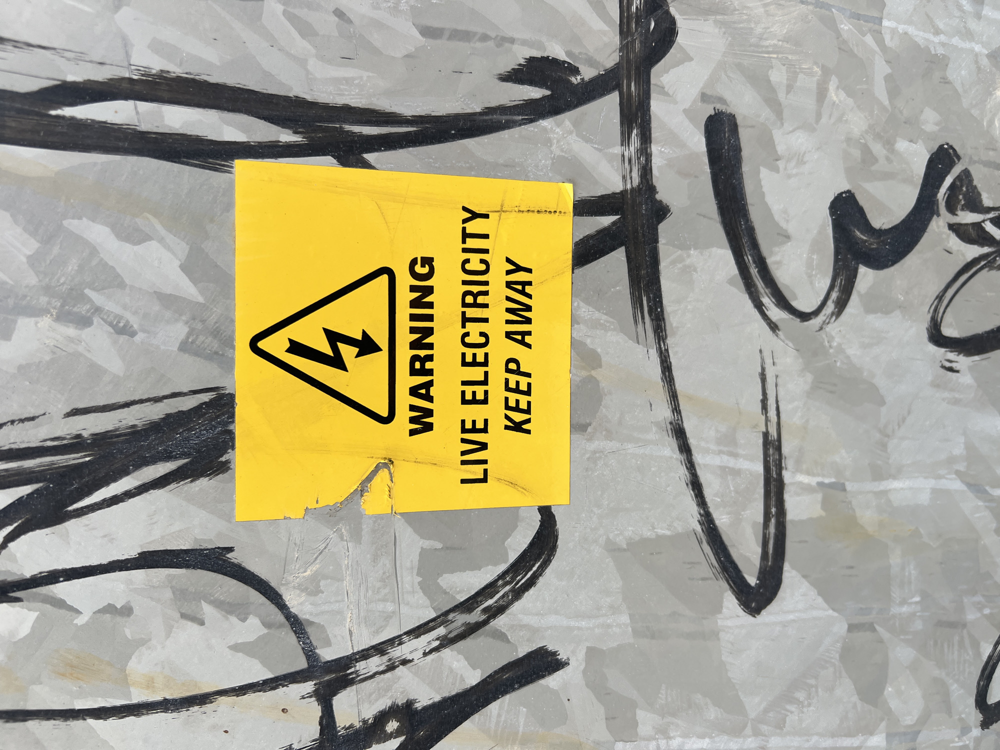 A yellow warning sign with a black lightning bolt symbol and the text "WARNING LIVE ELECTRICITY KEEP AWAY" is affixed to a metallic surface covered in black graffiti. The sign is torn and weathered. The metallic surface appears to be part of an outdoor utility box or enclosure. The graffiti consists of abstract black brushstrokes. The scene is likely an urban environment, possibly a street or alleyway in Balbriggan, Ireland, where such warning signs are common in areas with electrical infrastructure. No people are visible in the image.