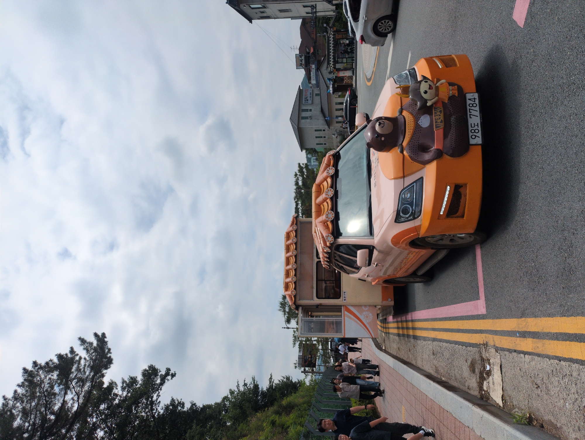 The image shows a novelty vehicle on a street in Gongju-si, South Korea. The vehicle, painted primarily in peach and orange, has decorations including roof-mounted ornaments resembling bread rolls, and character figures attached to the front. A license plate reads "98도 7784". Several people are standing and sitting near the sidewalk beside the vehicle. The street has pink and yellow painted lines. Buildings can be seen in the background. The sky is overcast. Vegetation is also visible on the left side of the image.