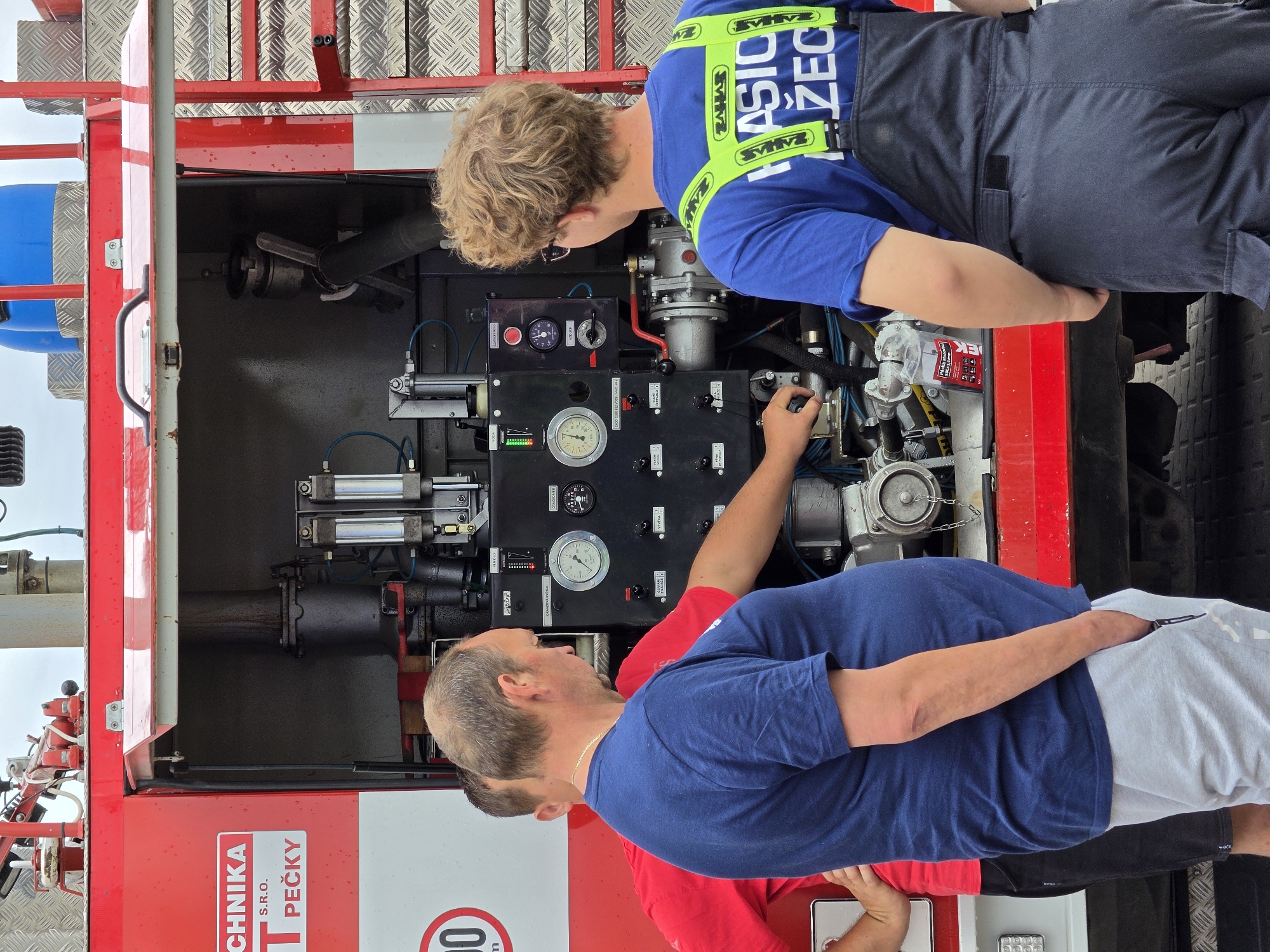 Two individuals are examining the controls of a fire truck. The person on the left wears a blue shirt and light grey athletic pants. The person on the right has curly blond hair, glasses and wears a blue shirt with the word "ZAHASICIO ŽEC" on it. The fire truck has the words "TECHNIKA PEČKY S.R.O." on its body. The fire truck controls include a gauge labeled "OTÁČKOMER" and a warning message "CHOO ČERP BEZ VODY MAX 60". The scene is in Mlčechvosty, Czechia.