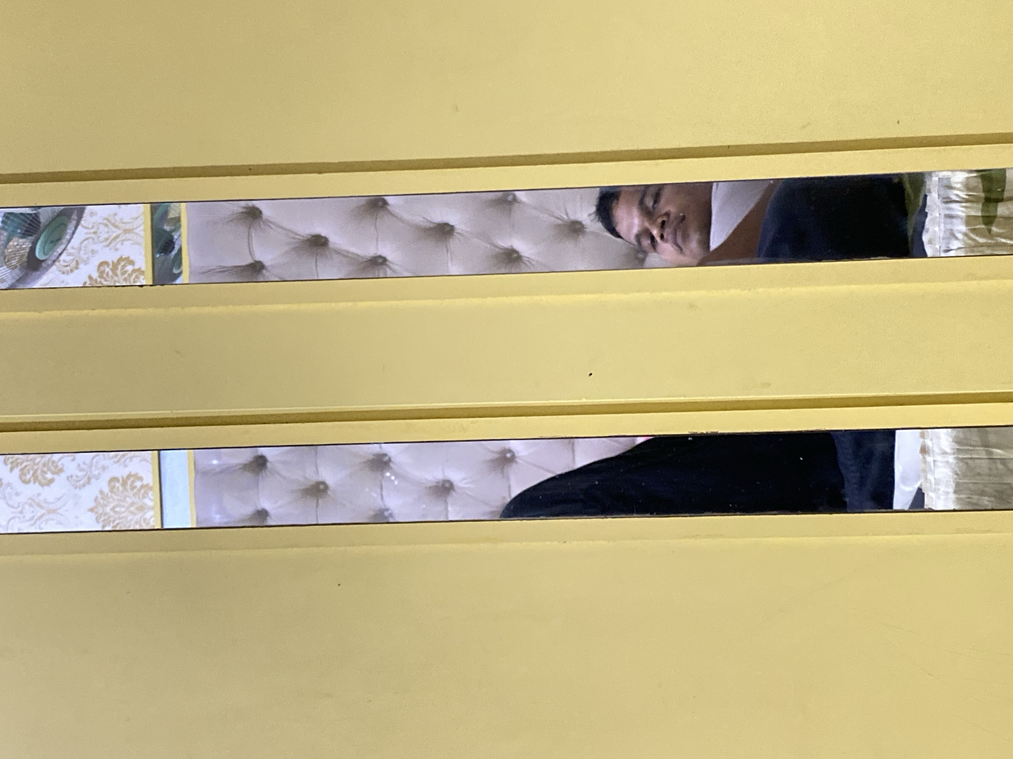 The image shows a close-up, vertical view of a yellow wall with two narrow, vertical openings. Through these openings, reflections of a tufted, upholstered surface, possibly a headboard or wall paneling, are visible. In the right opening, the reflection of a man's face is visible, looking upwards. He is wearing a white shirt and appears to be lying down or reclining. In the left opening, a dark garment, possibly clothing, is reflected. The lighting appears to be indoor. The exact location is Unknown, Unknown.