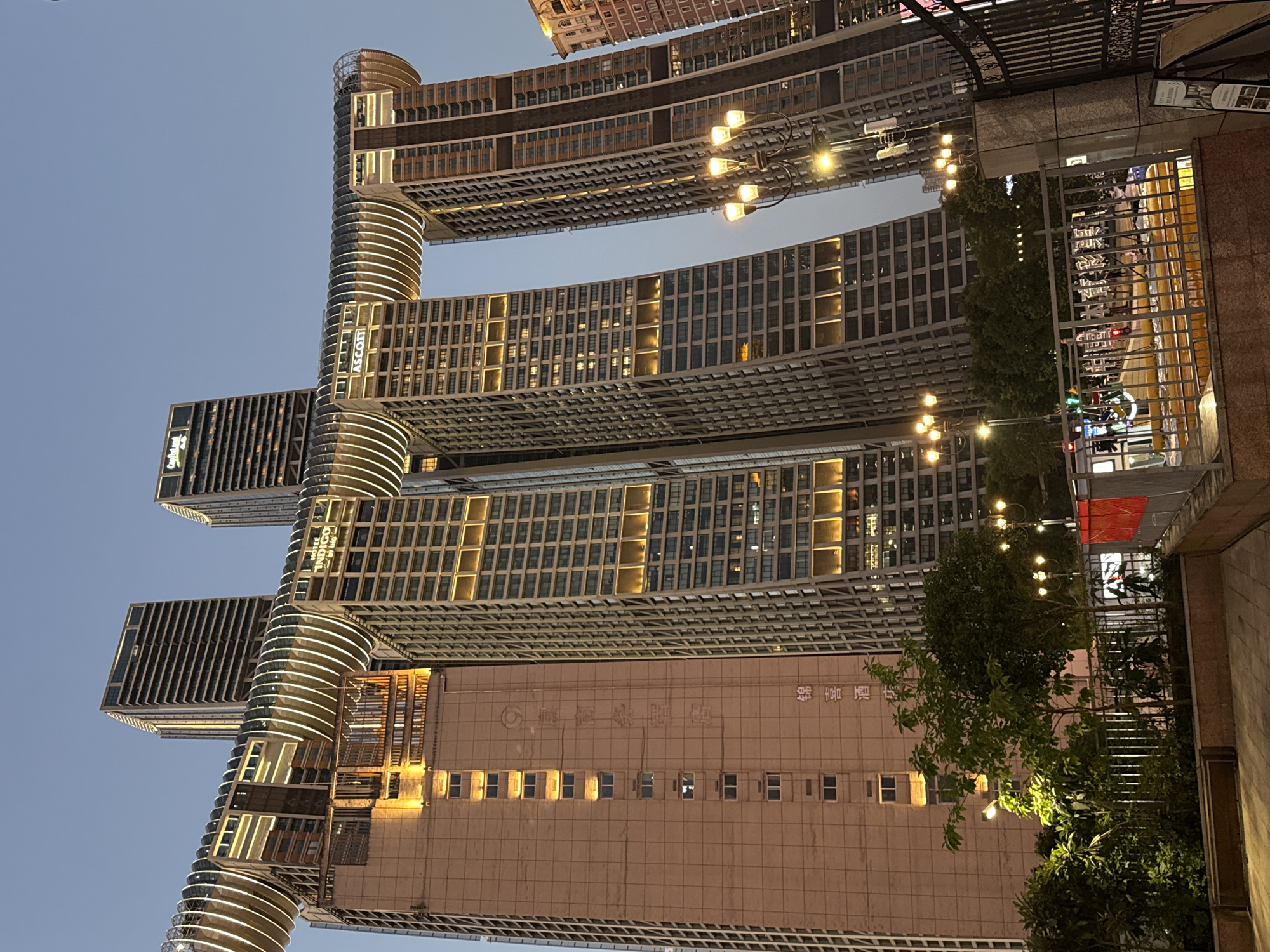 The image shows the Raffles City Chongqing complex, a mixed-use skyscraper complex located in the Yuzhong District of Chongqing, China. The complex consists of eight interconnected towers, with a unique horizontal skybridge that spans across four of the towers. The architecture is modern and striking, with the towers featuring a distinctive, curved design. The skybridge is illuminated with a series of white lights, creating a dramatic effect against the twilight sky.
The towers are clad in glass and metal, with visible windows and balconies. Some of the towers have signage visible, including "HOTEL INDIGO" and "ASCOTT". The complex is situated along the Yangtze River, and the surrounding urban landscape is visible in the background, with other buildings and city lights. In the foreground, there are trees, streetlights, and a metal fence, suggesting a public area or pedestrian walkway. The overall scene captures the impressive scale and innovative design of the Raffles City Chongqing complex.