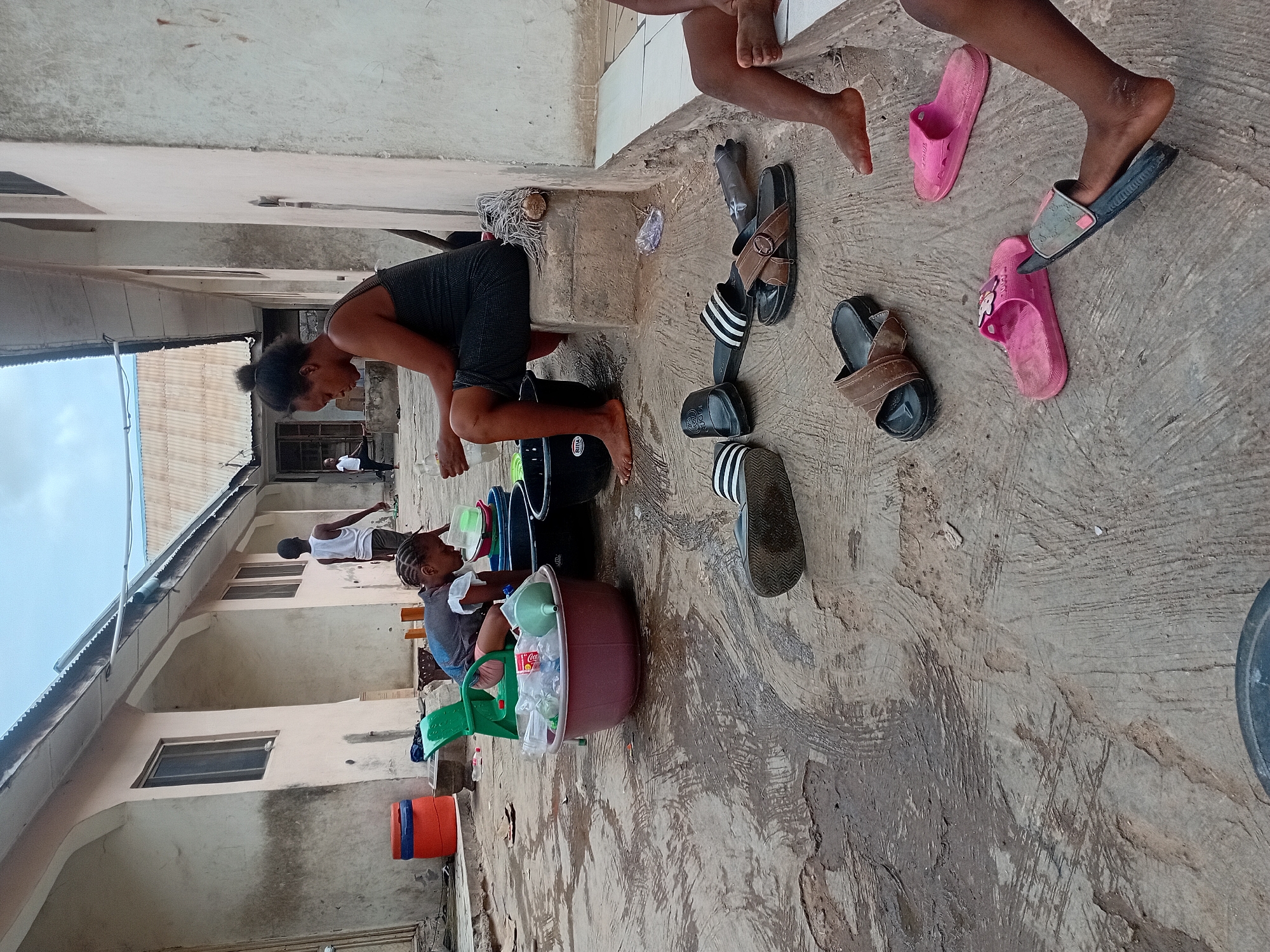 This image, rotated 90 degrees counter-clockwise, depicts an outdoor scene in what appears to be a residential area, likely in Lokoja, Nigeria, given the context. The time of day is daytime, with soft natural light suggesting either an overcast sky or early morning/late afternoon.
The primary subjects are a woman and a child engaged in an activity involving water. The woman, wearing a dark sleeveless top and shorts, is bent over a large dark basin or bucket, appearing to be pouring or handling liquids. Her bare feet are visible on the wet, concrete ground. Next to her, a child with braided hair, dressed in a light top, is seated on a green plastic stool and also interacting with containers.
Numerous objects are present, indicating a routine household activity. These include several buckets and basins of various sizes, a green watering-can-like container, a funnel, and plastic bottles, some containing liquid and others seemingly empty or discarded. A black bucket features the "RITA" logo, and a plastic bottle inside a basin shows the "Coca-Cola" branding. Scattered around are multiple pairs of slippers and sandals in different styles (striped, brown, pink), along with the bare feet of several individuals, suggesting more people are present or nearby, possibly children. A red and blue cooler or large bucket is visible in the background.
The setting consists of a rough, grey concrete ground that is visibly wet in patches, indicating the use of water. In the background, multi-story buildings with light-colored, possibly stuccoed or painted walls and windows are visible. A corrugated metal roof extends across the upper part of the image, contrasting with the sky which appears bright but lacks strong sun. In the deeper background, another person wearing a white top is faintly discernible. The overall scene conveys a sense of daily life and communal activity in an outdoor domestic space.