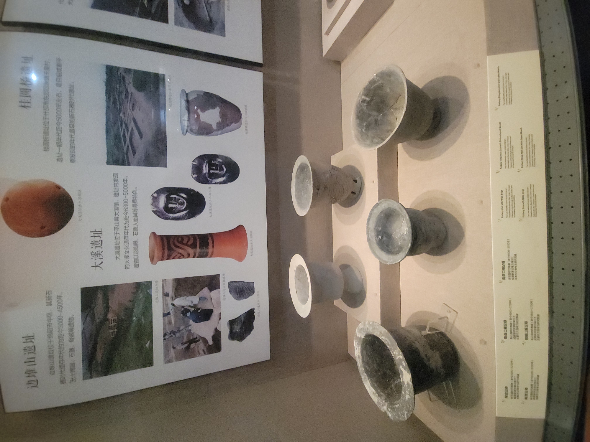 The image displays an exhibit of ancient Chinese artifacts, with text in Chinese. The exhibit is divided into sections, each focusing on a specific archaeological site. One section is titled "边堆山遗址" (Bian Duishan Site), located in the city of Qingyang District, China. The text indicates this site dates back to the Neolithic period, specifically 5000-4500 years ago, and has yielded pottery, stone tools, and bone artifacts. Another section is labeled "大溪遗址" (Daxi Site), also described as dating to the Neolithic period, approximately 6300-5000 years ago, and features pottery and human face-shaped artifacts. The third main section is "桂圆桥遗址" (Guiyuanqiao Site), which is stated to be approximately 5000 years old and is considered the earliest Neolithic site discovered in the region.
In the foreground, several pottery vessels are displayed on a light-colored platform. These vessels vary in size, shape, and color, with some appearing to be made of grey or dark clay, while others are lighter in hue. The background shows enlarged photographs or illustrations of the archaeological sites and some of the artifacts recovered from them, including pottery, stone tools, and what appear to be carved human faces. The overall presentation suggests a museum or exhibition setting.