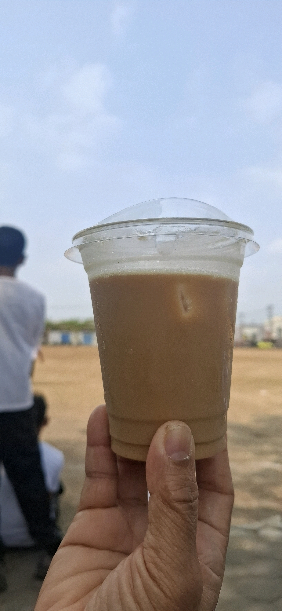 An outdoor, slightly low-angle, medium shot captures a hand holding a clear plastic cup filled with a light brown beverage, likely coffee or tea, with ice cubes visible. The cup has a domed, clear plastic lid. The background is blurred, showing an open, dusty area with a few individuals, one in a white shirt, standing in the distance. The sky above is a pale blue with sparse white clouds. The image appears to have been taken in Baturan, Indonesia, likely in an open field or recreational area.