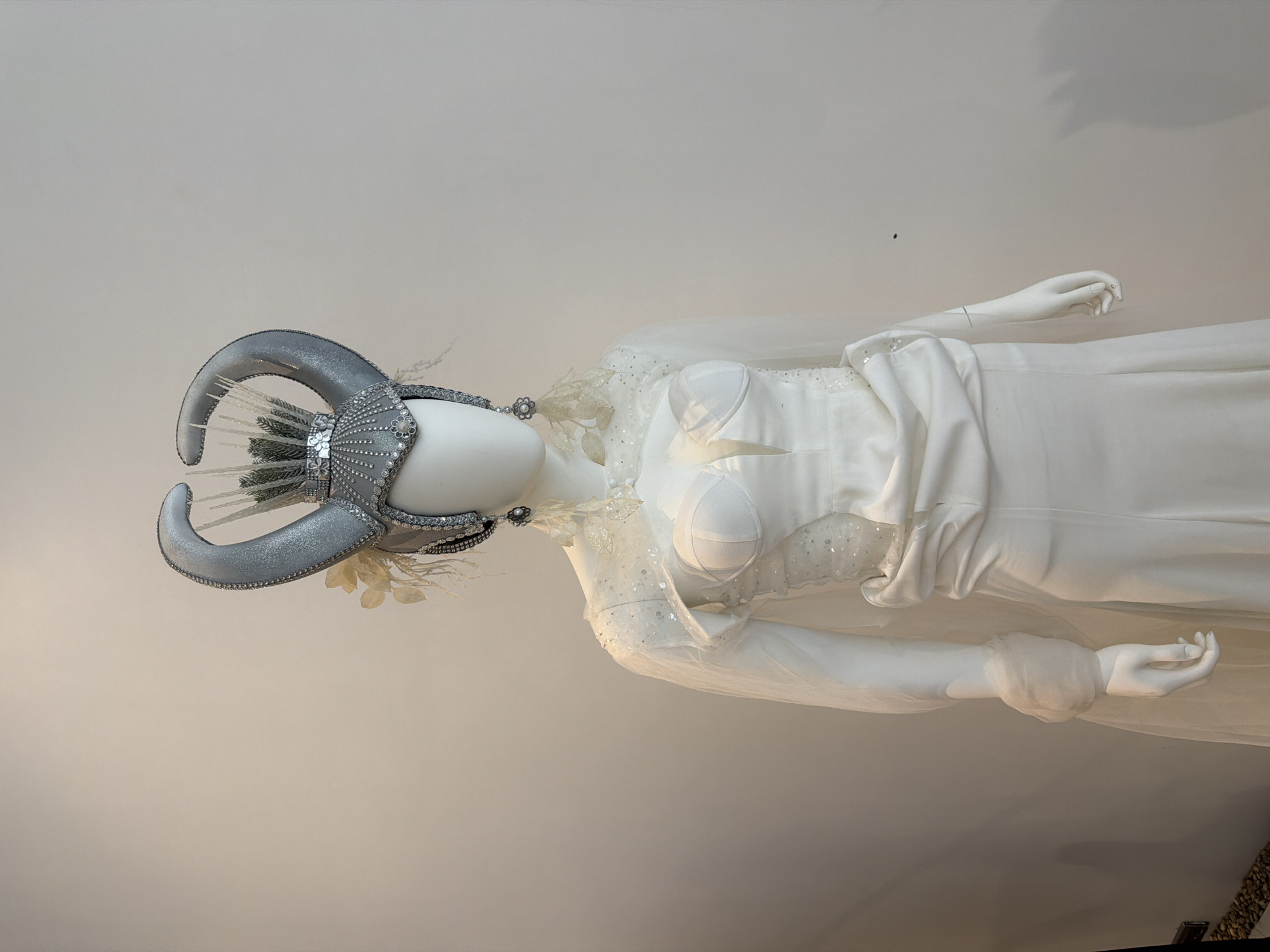 This is a full-body shot of a mannequin dressed in an elaborate white gown with a dramatic silver headdress. The headdress resembles horns adorned with rhinestones and greenery. The gown features a structured bodice with sheer, sequined shoulder straps and long, flowing sleeves. A draped skirt completes the look. The mannequin is positioned against a plain, light-colored wall, suggesting an indoor setting, possibly a boutique or exhibition space. There is no indication of the location being in Ko Samui District, Thailand.