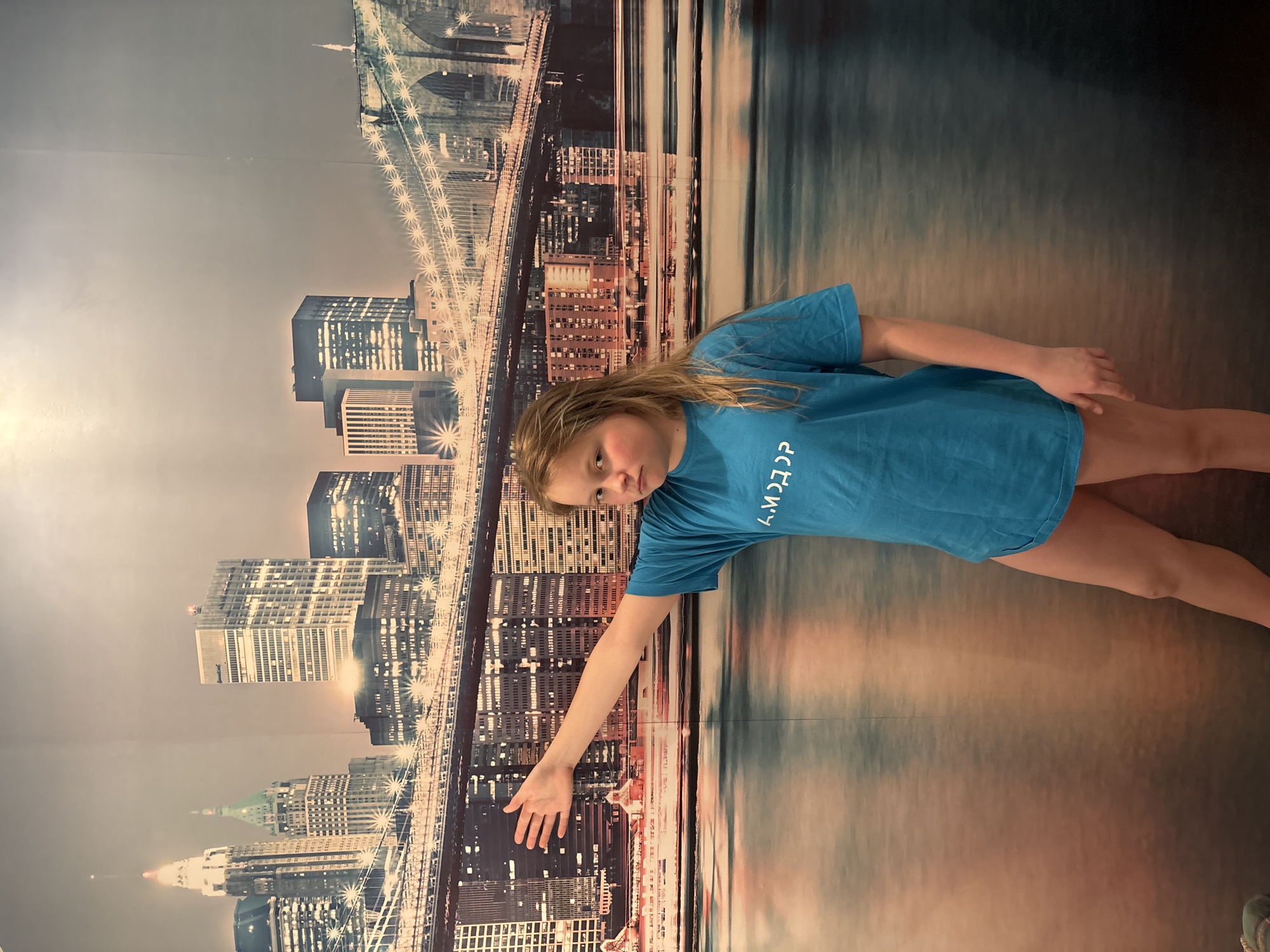 An indoor photograph, taken in Русский Вожой, Russia, features a young individual standing centered against a large mural depicting the illuminated New York City skyline at dusk, including the Brooklyn Bridge and various skyscrapers above a river. The individual, appearing to be a pre-teen, has light brown, shoulder-length hair and wears a loose, short-sleeved blue t-shirt with white Cyrillic text ("А.МОДЭР" visible) printed on the front. Light-colored upper thighs are visible below the t-shirt hem. The individual's right arm is extended horizontally to the side, palm open, pointing towards the left of the frame. Their gaze is directed towards the camera, with a neutral facial expression. Visible vertical seams in the mural indicate its manufactured nature. The scene is lit by interior artificial light.