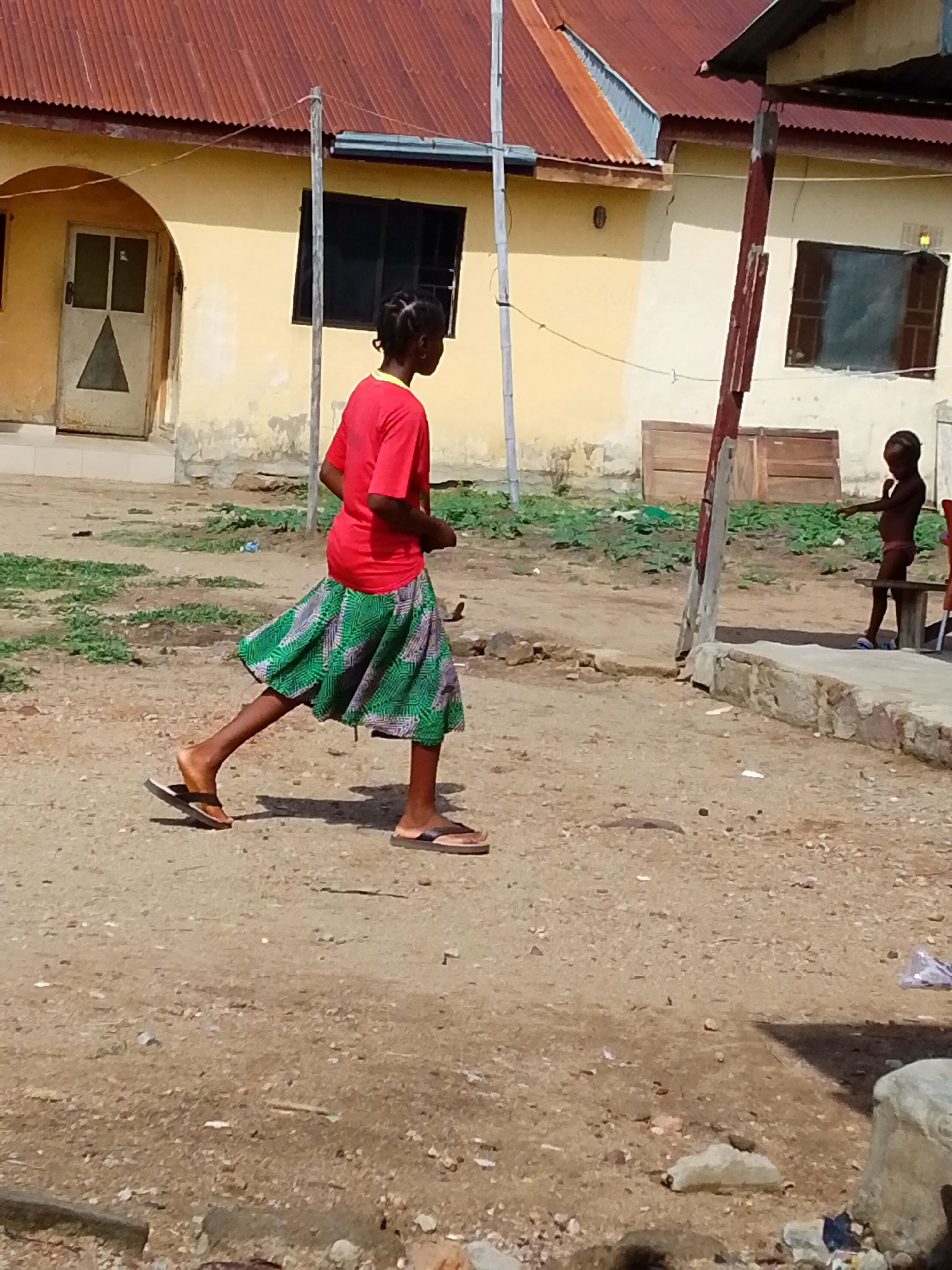 A sunny outdoor scene shows a young woman walking on a dirt ground. The woman is wearing a red shirt and a green and grey printed skirt. She is also wearing flip-flops. In the background are two buildings with corrugated iron roofs, one with a rusty brown roof, and the other, the roof is a light grey. The woman is walking past the buildings. Another young girl is partially visible next to a bench. The location appears to be Lokoja, Nigeria.