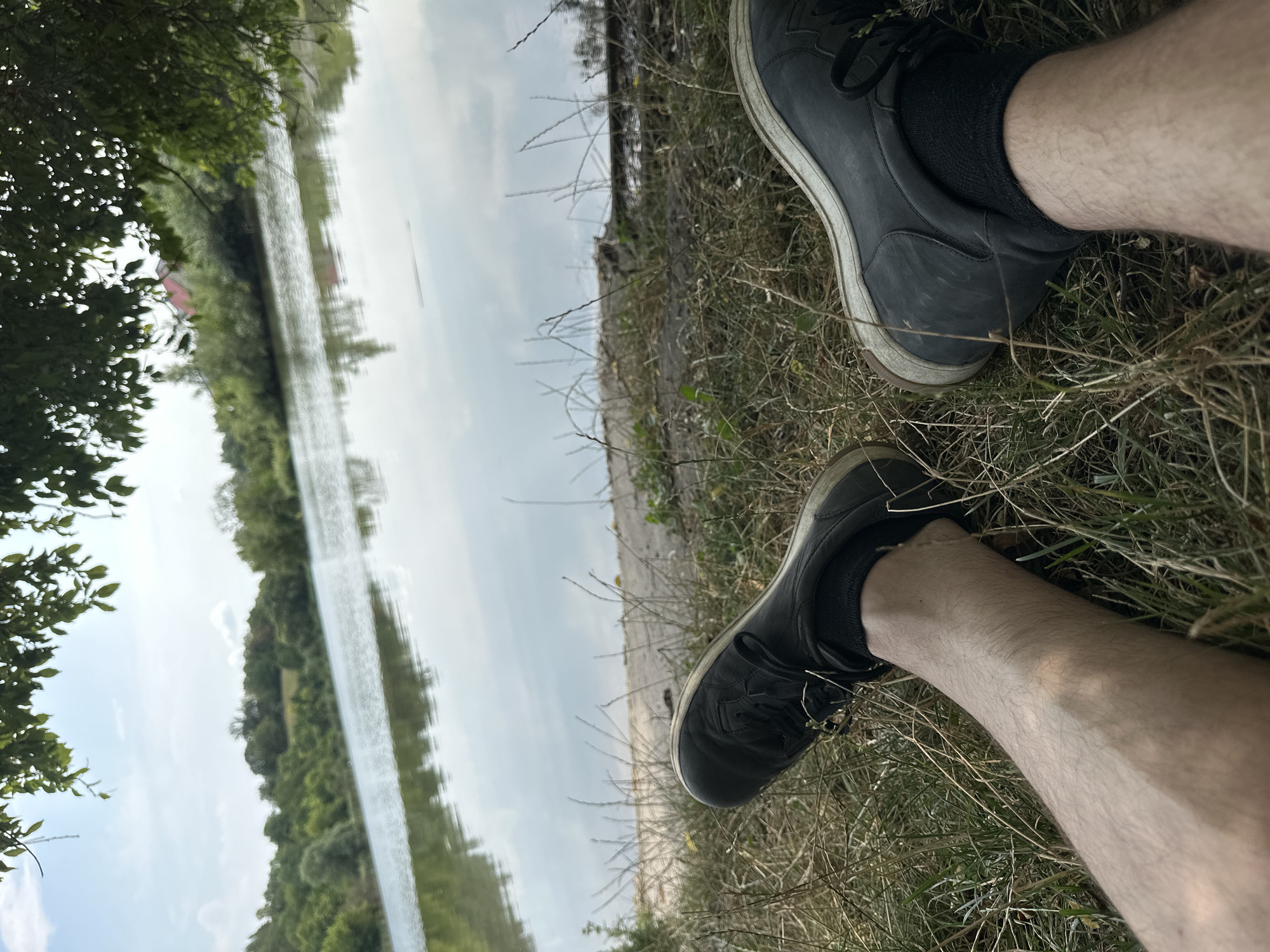 The image shows a pair of legs wearing black shoes and socks, resting on dry grass near a body of water. The background features trees and a cloudy sky, with a red-roofed building visible in the distance. The scene appears to be a natural outdoor setting, possibly a park or a lake shore, within or near the village of Makiv, Ukraine.